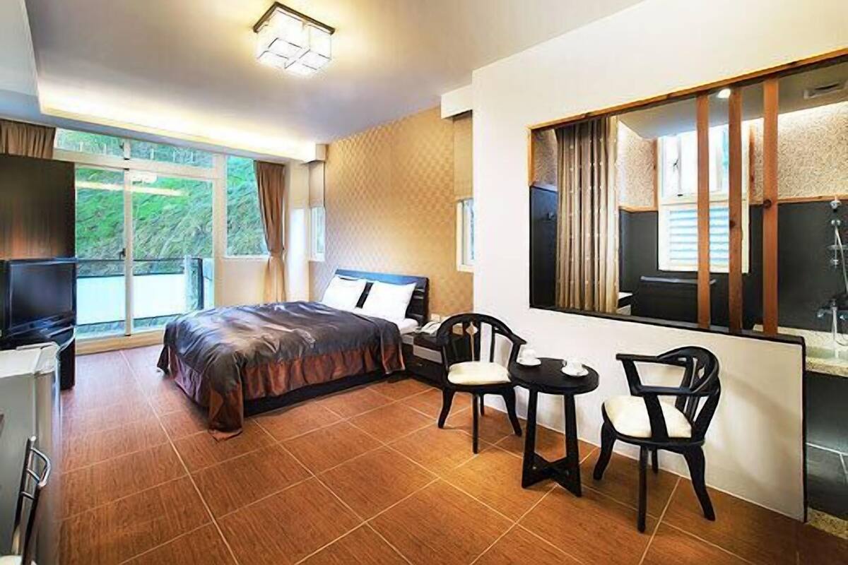 Quarto de hotel com cama, escrivaninha, duas cadeiras, televisão e uma janela grande com vista para áreas verdes.