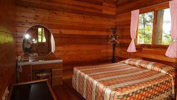 A wooden cabin room with a bed, a desk with a chair, a mirror, and a window with curtains.