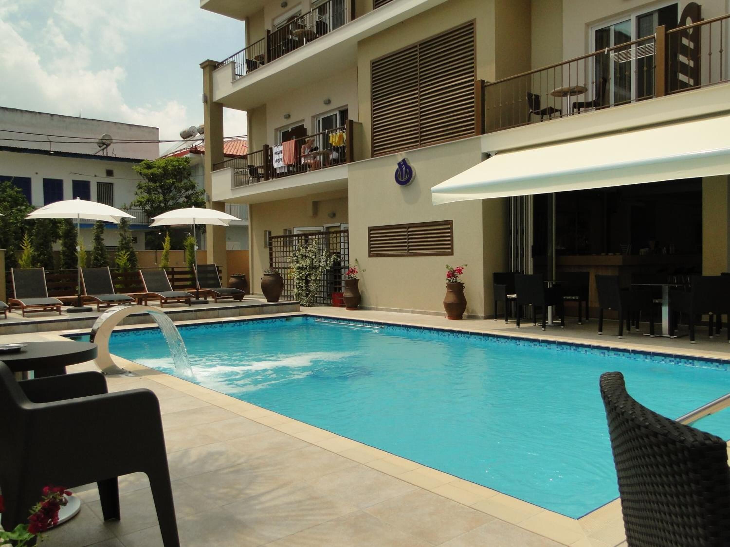 Una piscina con sillones de descanso y sombrillas, un área de estar cubierta y un edificio con balcones.