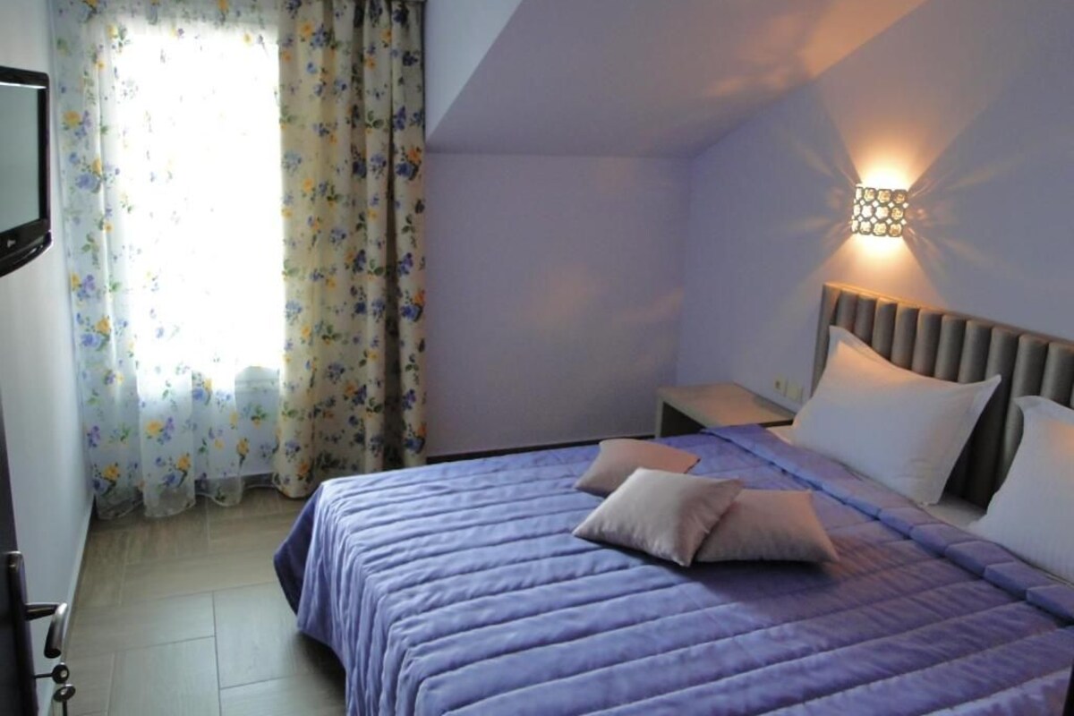 A bedroom with a bed, pillows, a nightstand, a wall-mounted light, and a window with curtains.