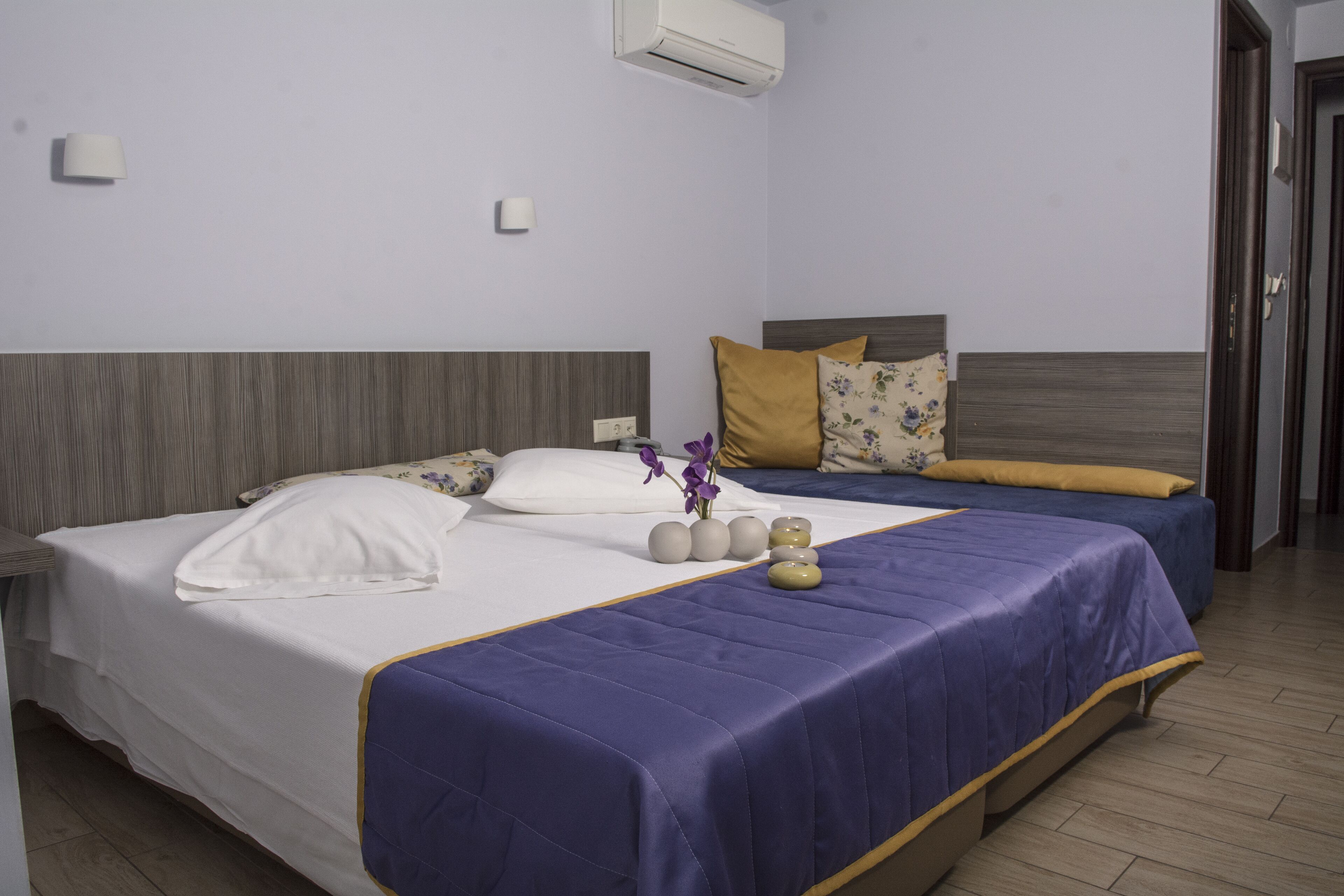 A hotel room with two beds, a wooden headboard, a purple bedspread, and a small vase with flowers.