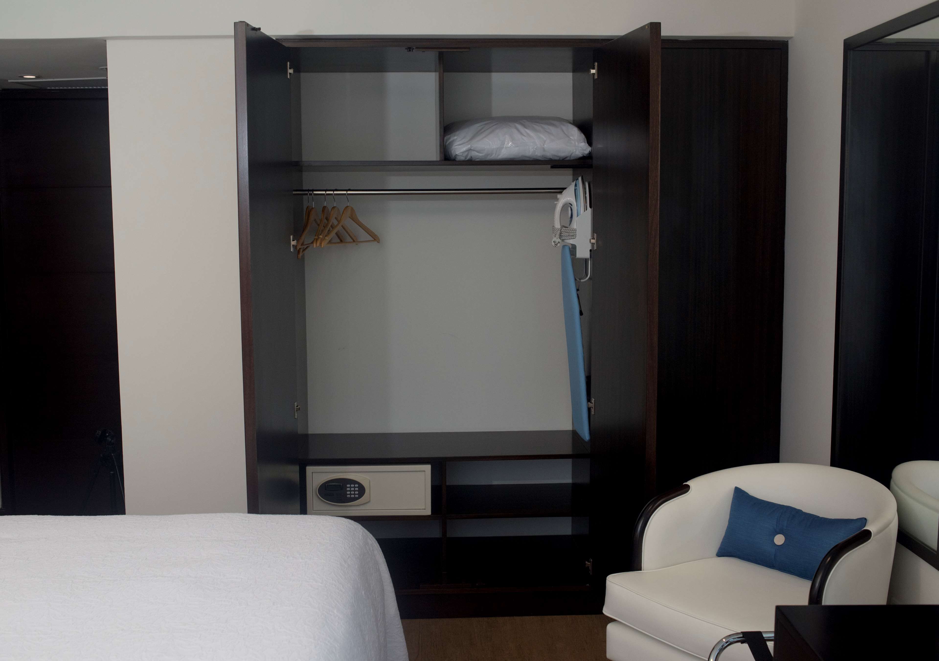 A hotel room with a bed, a white armchair with a blue pillow, a wardrobe with a hanging rack, and a small desk with a chair.