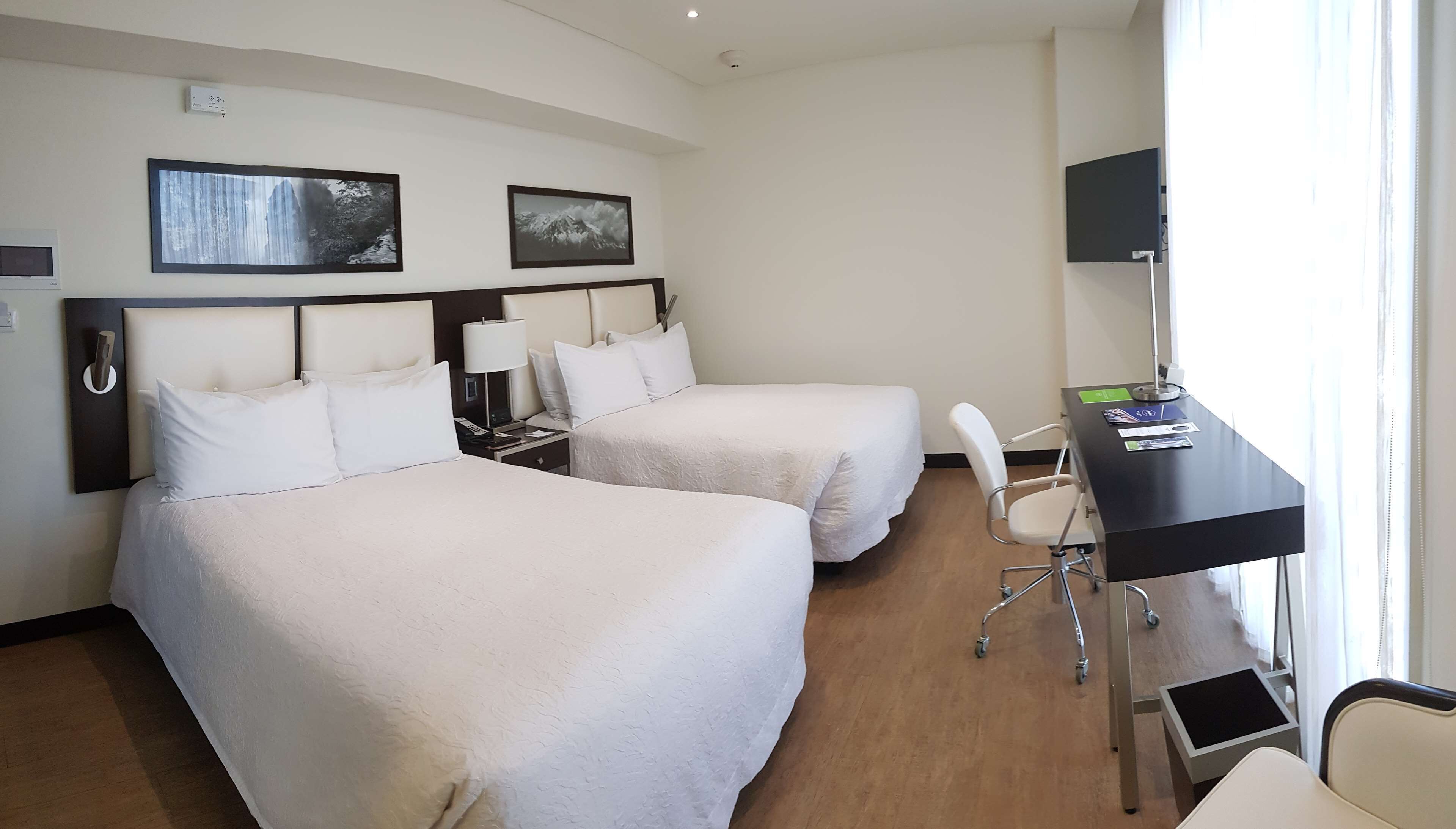 A hotel room with two beds, a desk with a chair, a TV, and artwork on the wall.