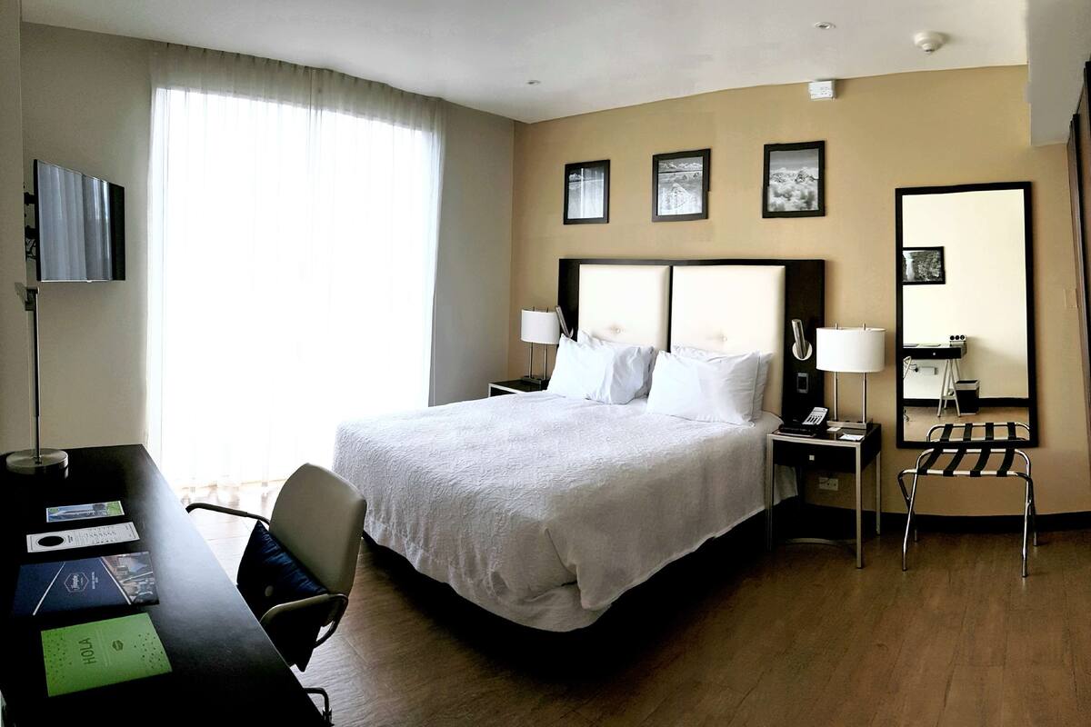 Ein Hotelzimmer mit einem großen Bett, einem Schreibtisch mit Stuhl, einer Schreibtischlampe, einem Telefon und einem Spiegel. An der Wand hängen drei gerahmte Bilder.