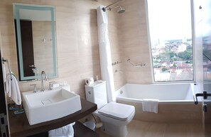 A modern bathroom with a large sink, toilet, and bathtub, a mirror, and a view of the city through a large window. - Hampton by Hilton Santa Cruz/Equipetrol, Bolivia (Santa Cruz)