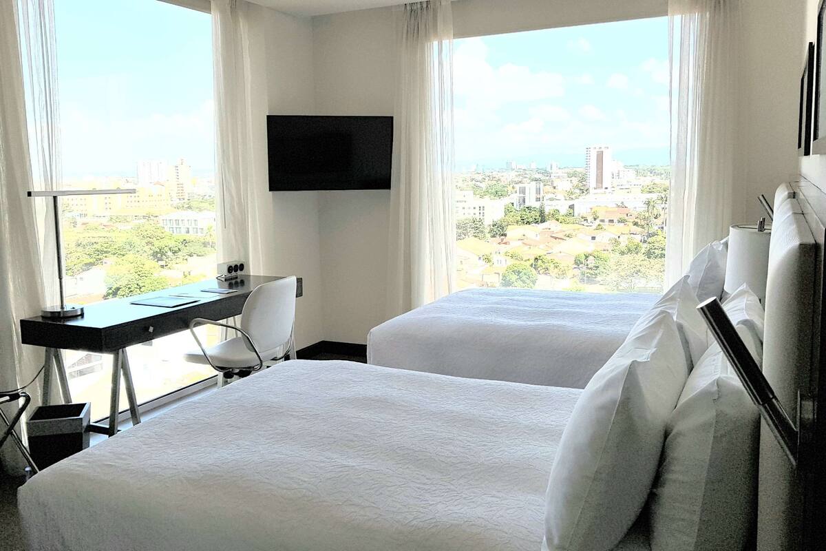 Ein Hotelzimmer mit zwei Betten, einem Schreibtisch, einem Sessel und einem Fernseher, mit Blick auf die Stadt.