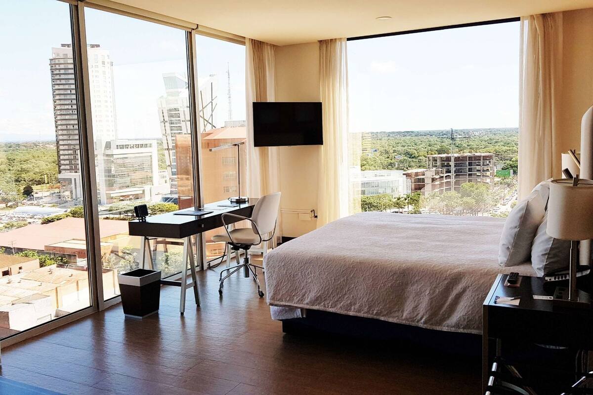 Ein Hotelzimmer mit einem großen Bett, einem Schreibtisch mit Stuhl, einem an der Wand montierten Fernseher und Blick auf die Stadt.