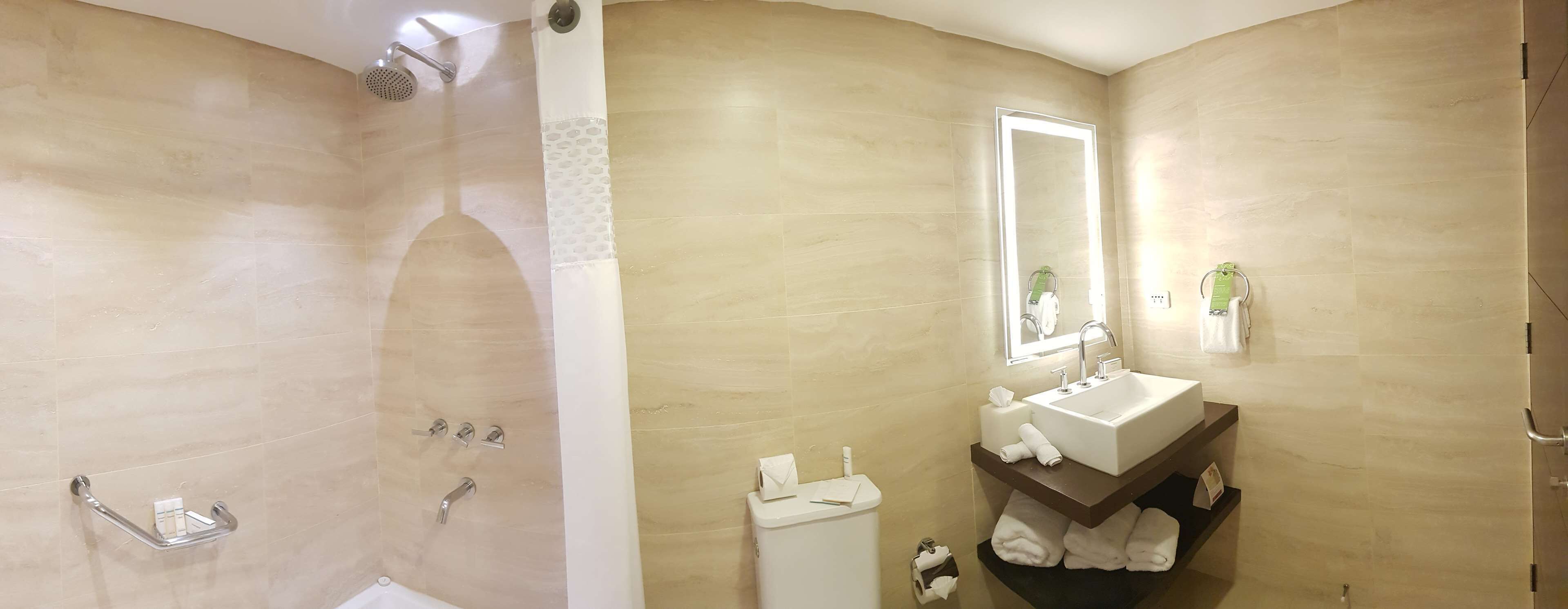 Baño con baldosas color beige, ducha con cabezal de ducha manual, inodoro y lavamanos con espejo encima.