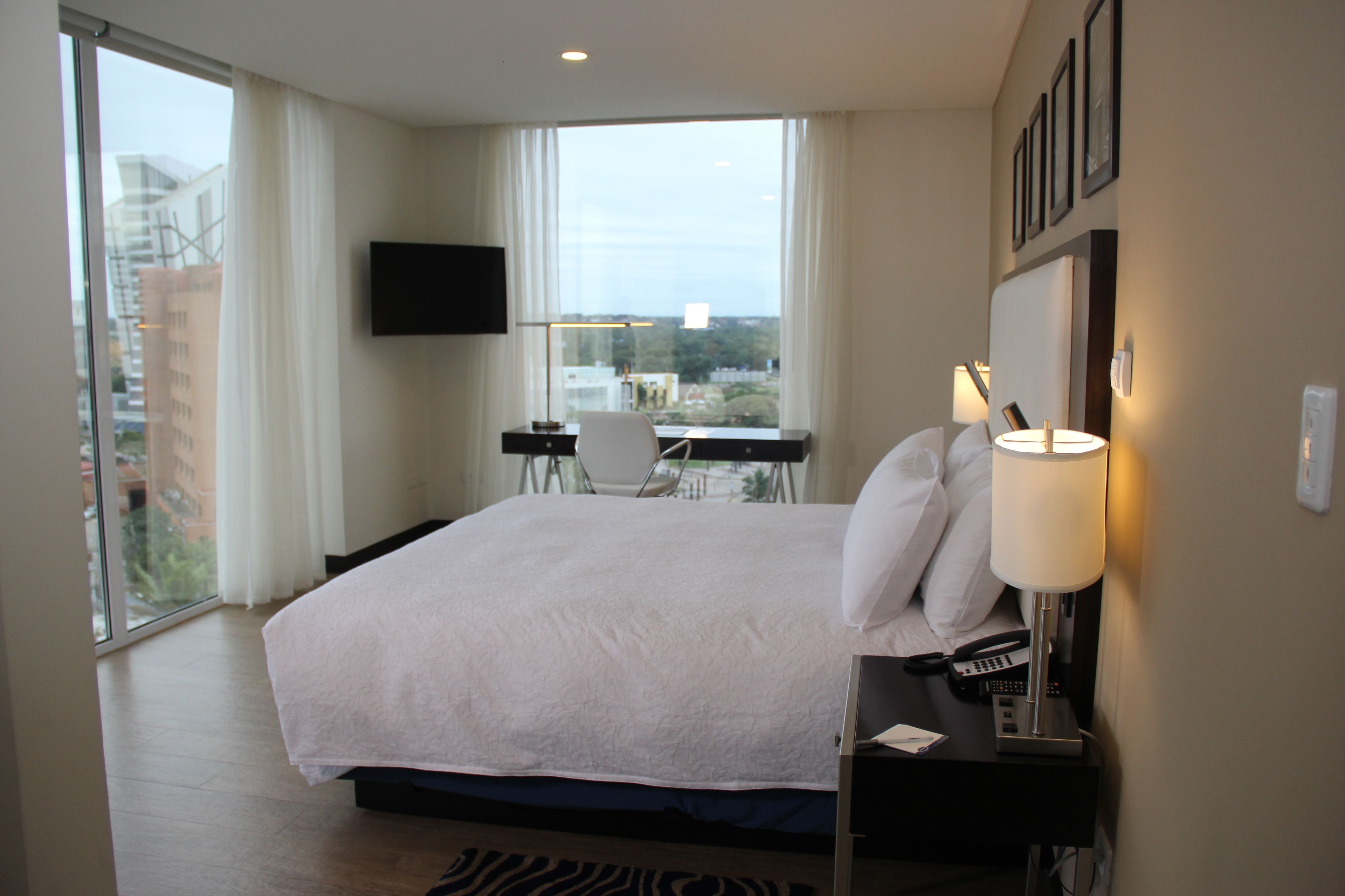 A hotel room with a large bed, a desk with a chair, a flat-screen TV mounted on the wall, and a window offering a city view.