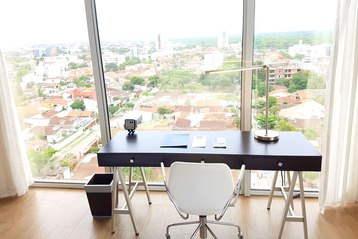 Ein moderner Schreibtisch mit Laptop, Lampe und Stuhl, vor einem großen Fenster mit Blick auf eine Stadtlandschaft.
