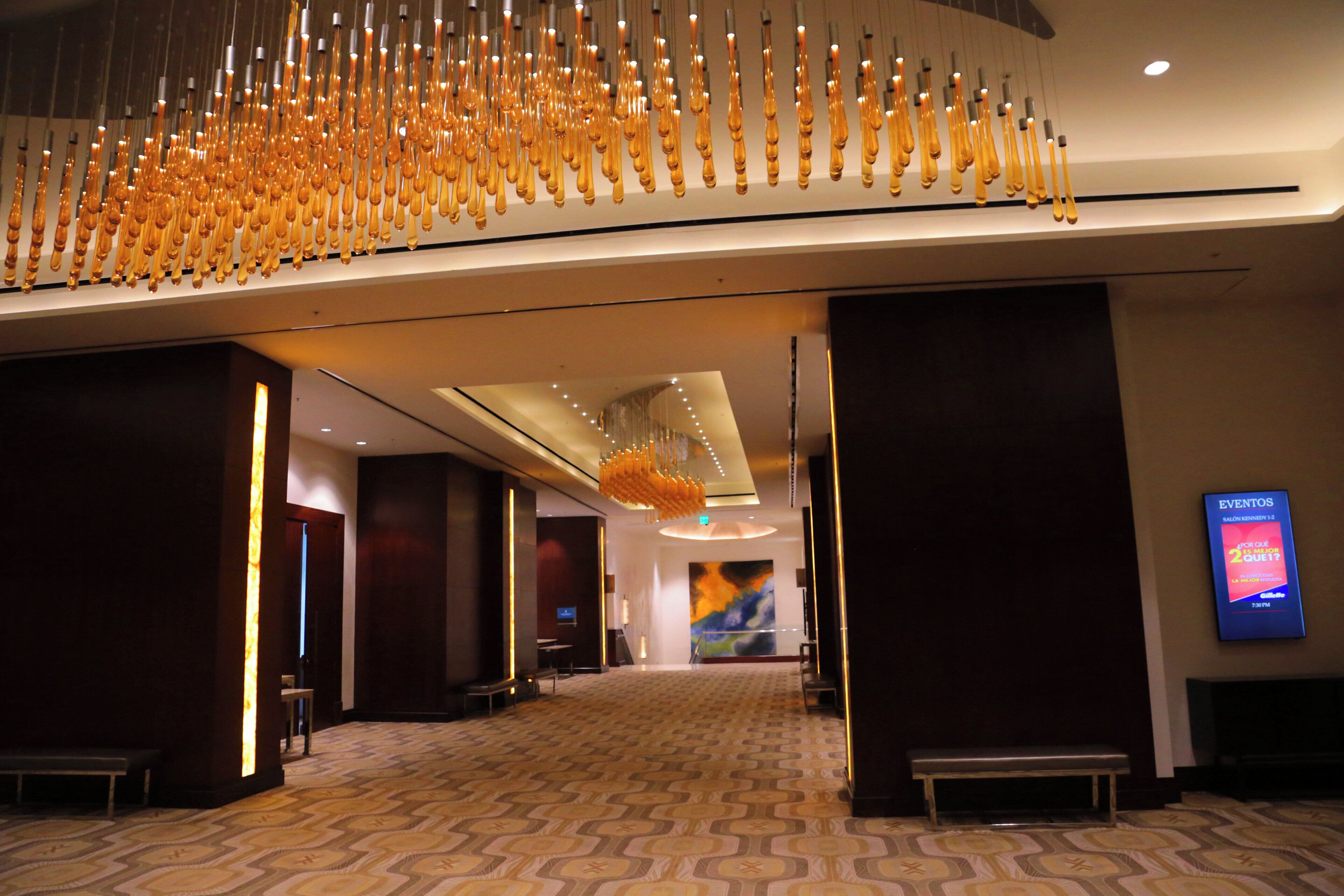 A spacious hotel lobby with a patterned carpet, a modern chandelier, and a digital event board.
