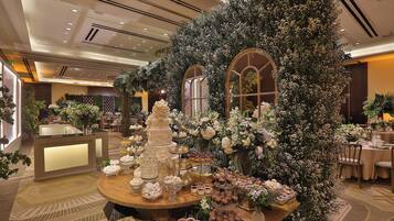 Eine luxuriöse Hotellobby mit einem runden Tisch in der Mitte, der eine Auswahl an Desserts und Kuchen präsentiert, umgeben von aufwendigen Blumenarrangements und stilvollem Mobiliar.
