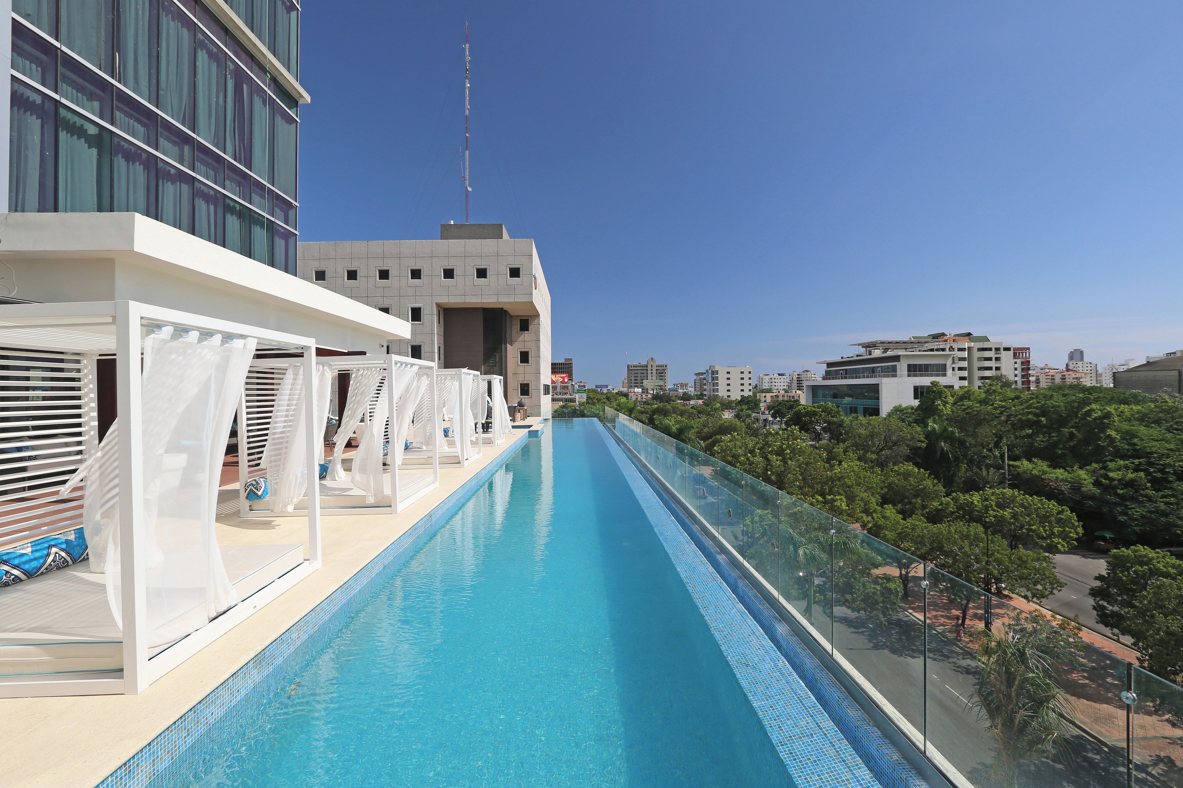 Una piscina larga y estrecha con una barrera de cristal en un lado, rodeada de sillones de descanso y sombrillas, con edificios al fondo.