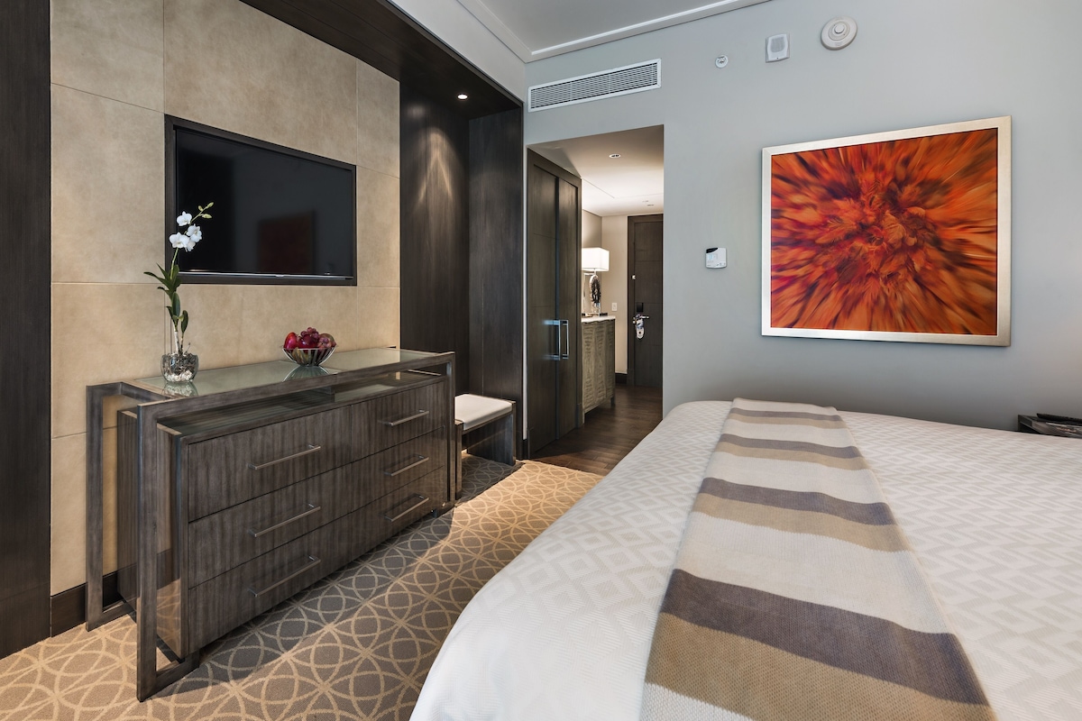 A modern hotel room with a large bed, a dark wooden dresser, a flat-screen TV, and a vibrant abstract painting on the wall.