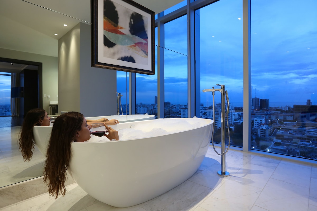 A modern bathroom with a large bathtub, a city view, and a framed artwork on the wall.