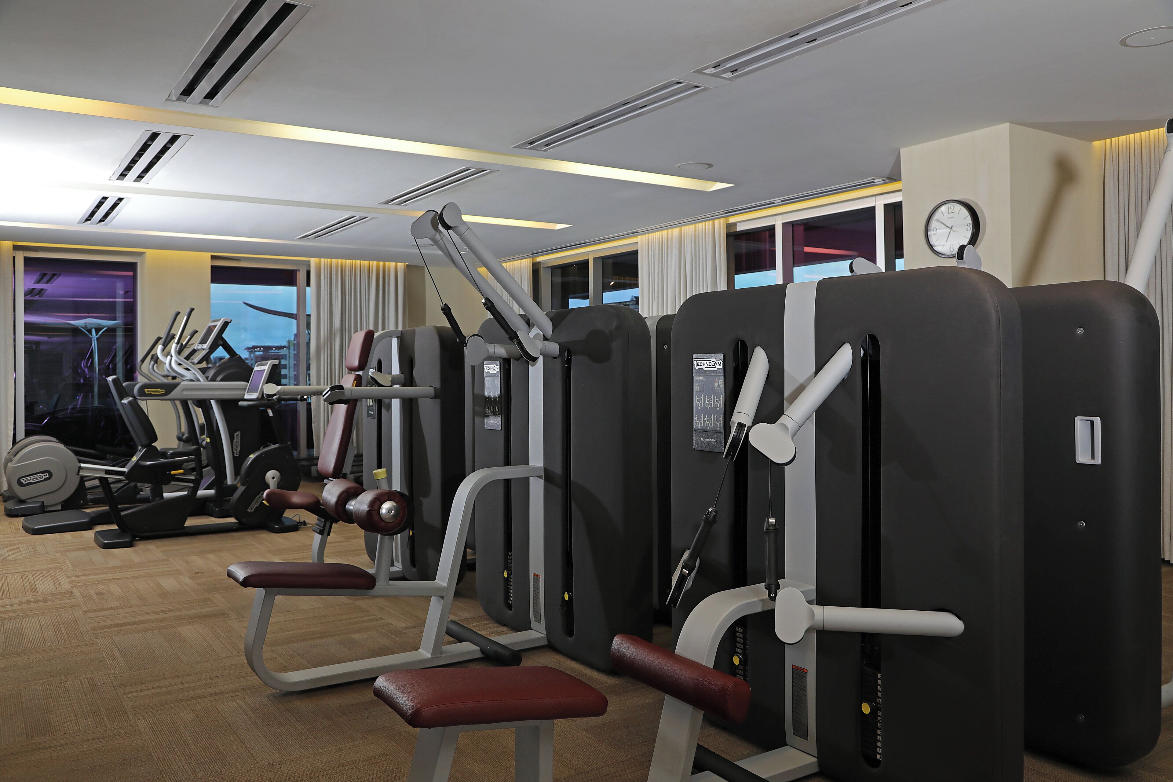 A gym with various exercise machines, a clock on the wall, and large windows with curtains.