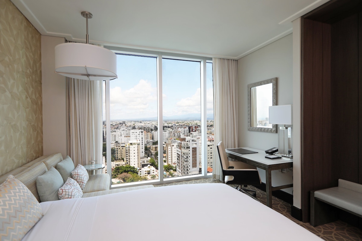 A hotel room with a large bed, a desk, a chair, a lamp, and a view of the city through large windows.