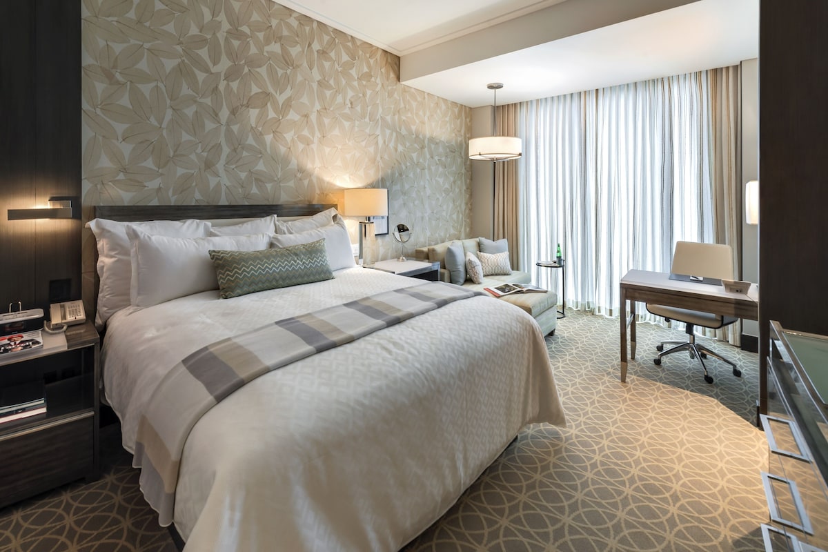A hotel room with a large bed, a desk, a chair, a lamp, and a telephone.