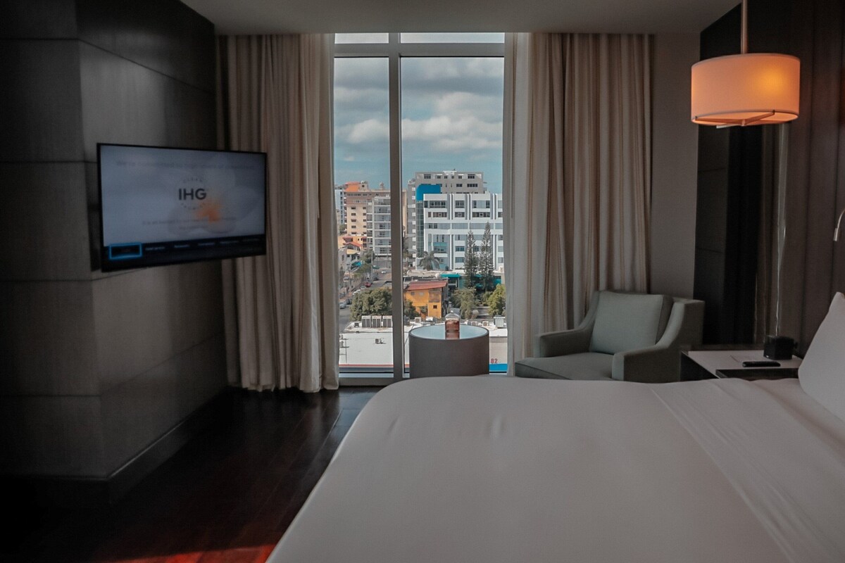 Een hotelkamer met een groot bed, een aan de muur bevestigde televisie, een stoel, een klein tafeltje en uitzicht op de stad door het raam.