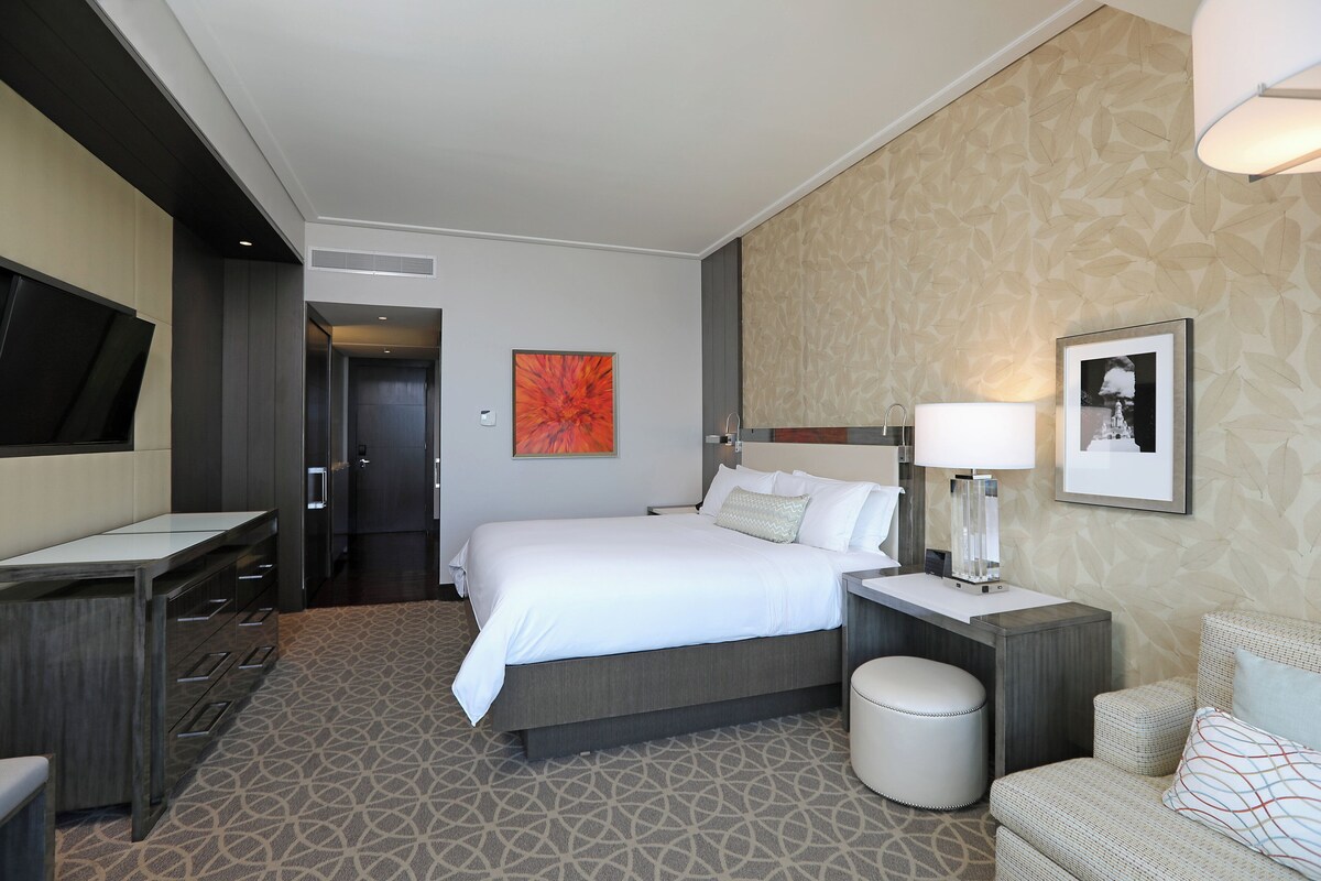 Een hotelkamer met een bed, nachtkastje, lamp, stoel en een televisie aan de muur.