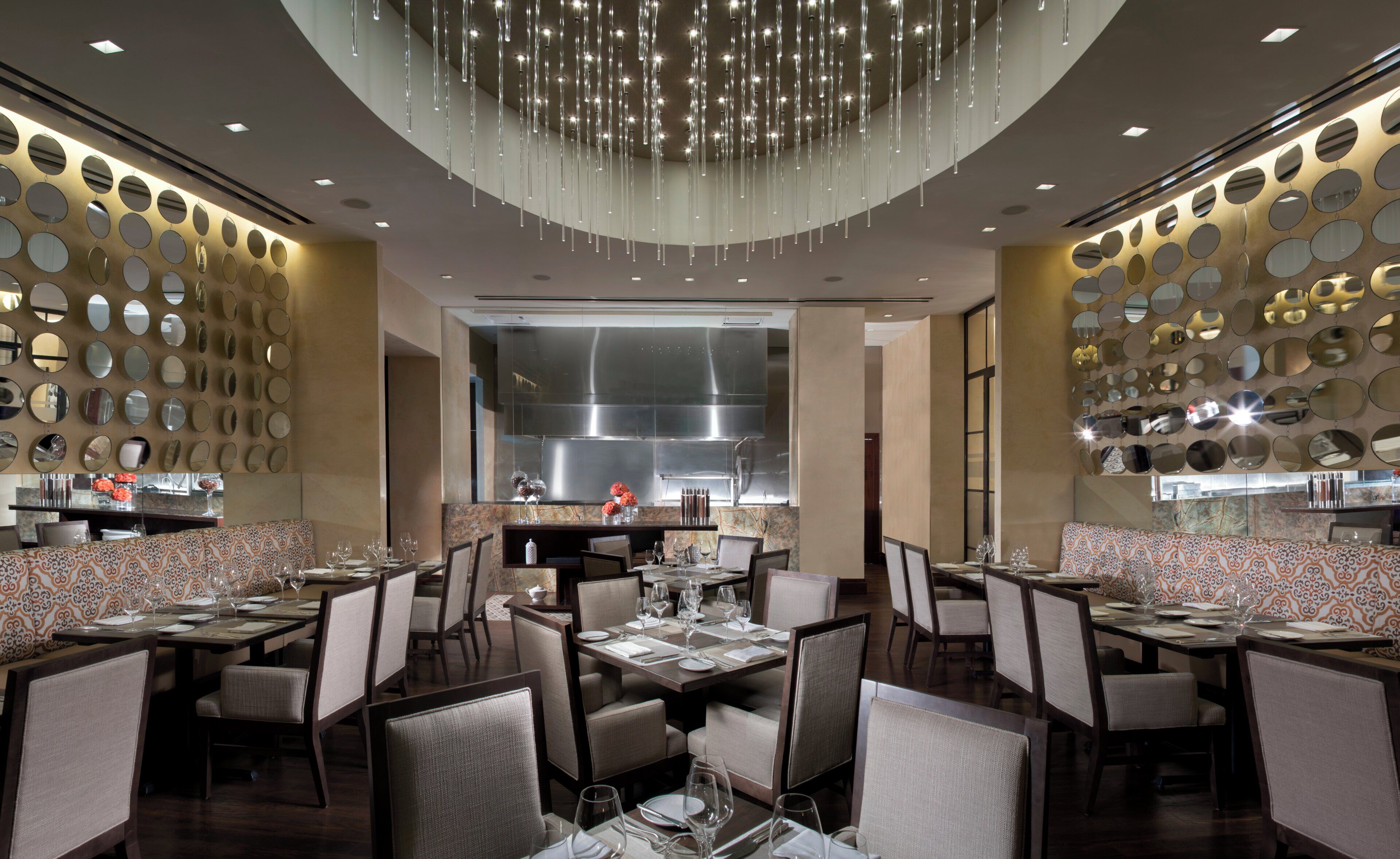 A restaurant interior with multiple dining tables, chairs, a large central kitchen, and a decorative ceiling light fixture.
