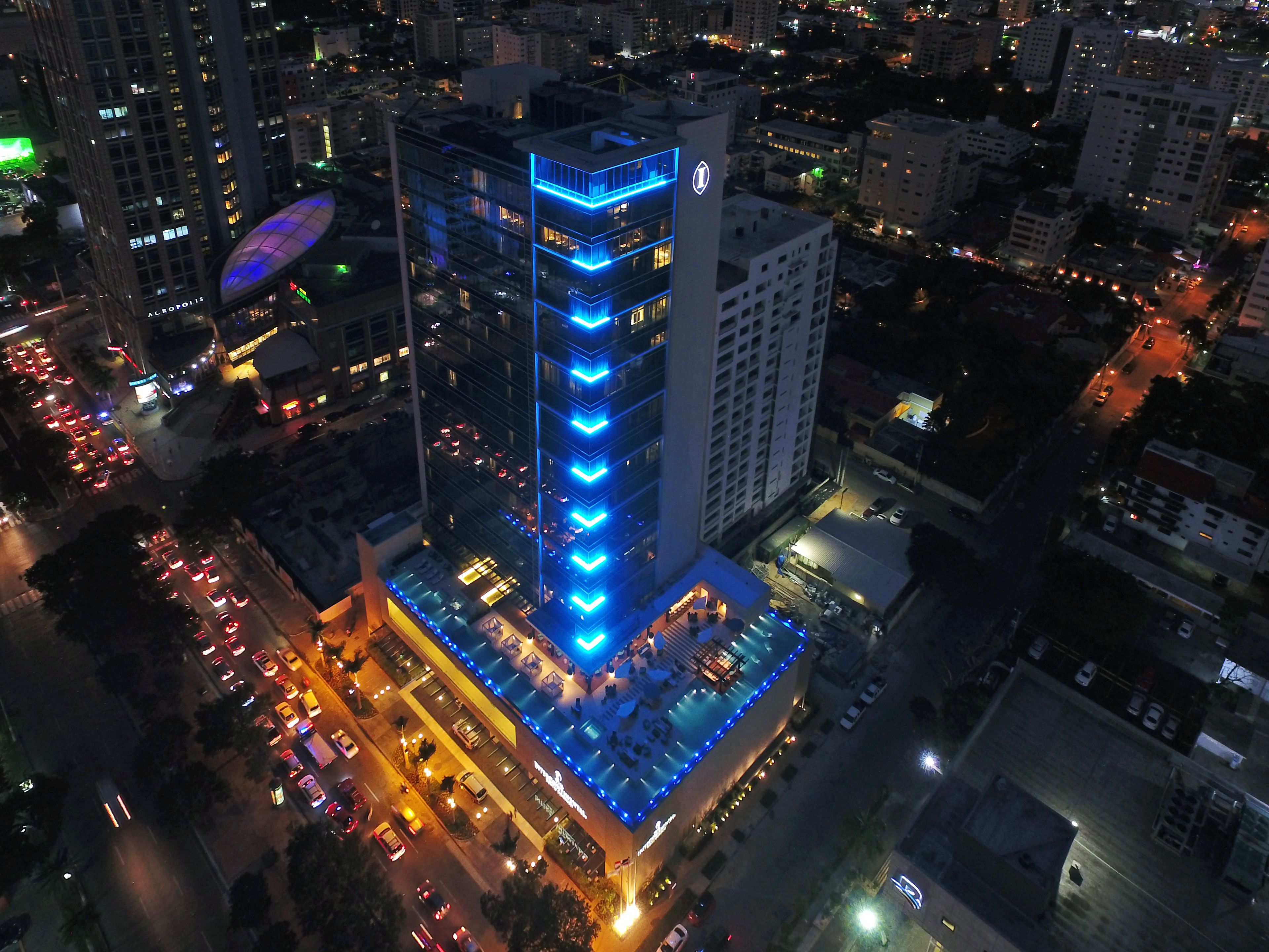 Un hotel de gran altura con una fachada distintiva iluminada en azul, un área de piscina en la azotea y un estacionamiento.