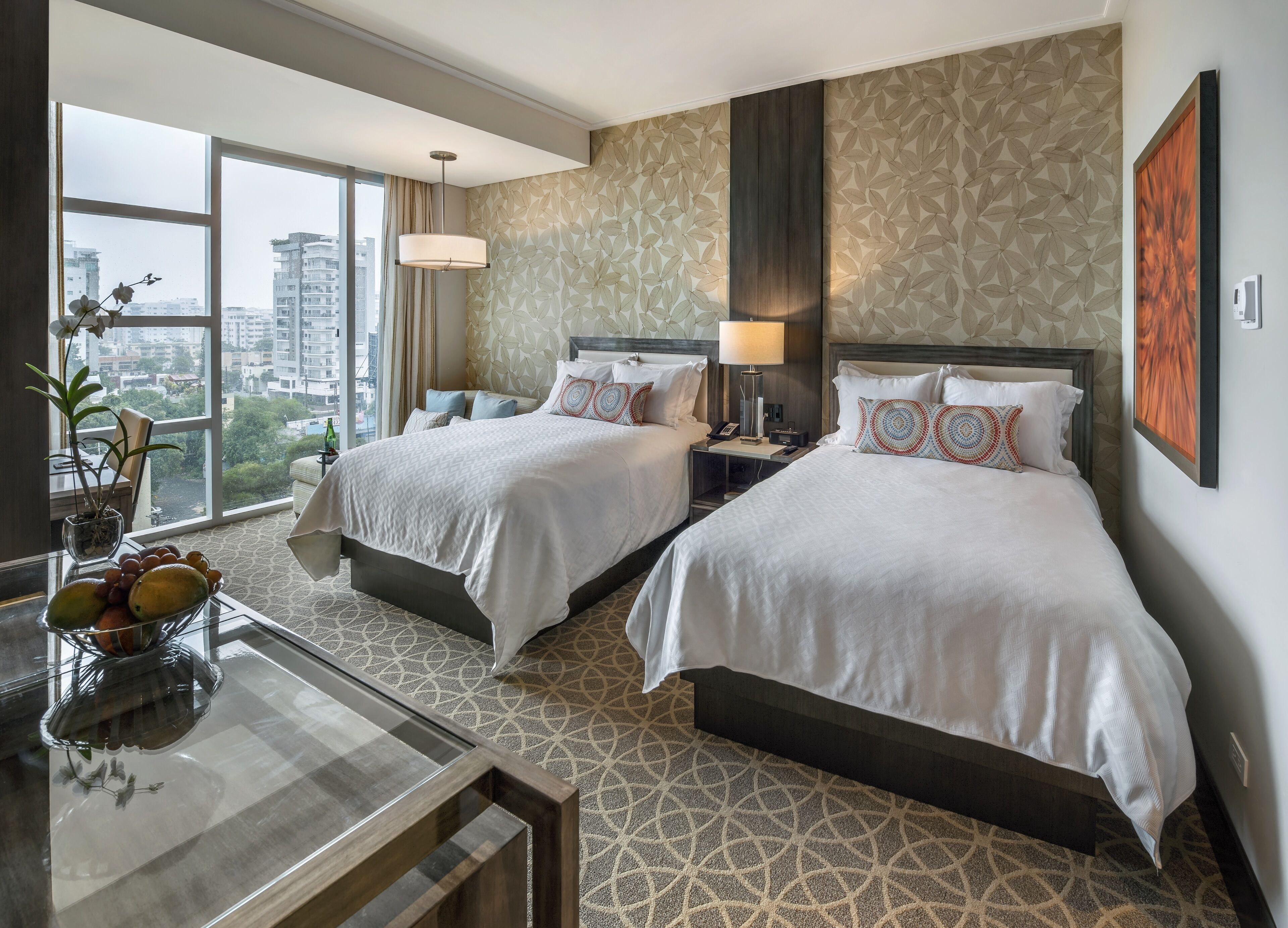 Habitación de hotel con dos camas, una mesa de comedor con fruta, vistas a la ciudad y una alfombra estampada.