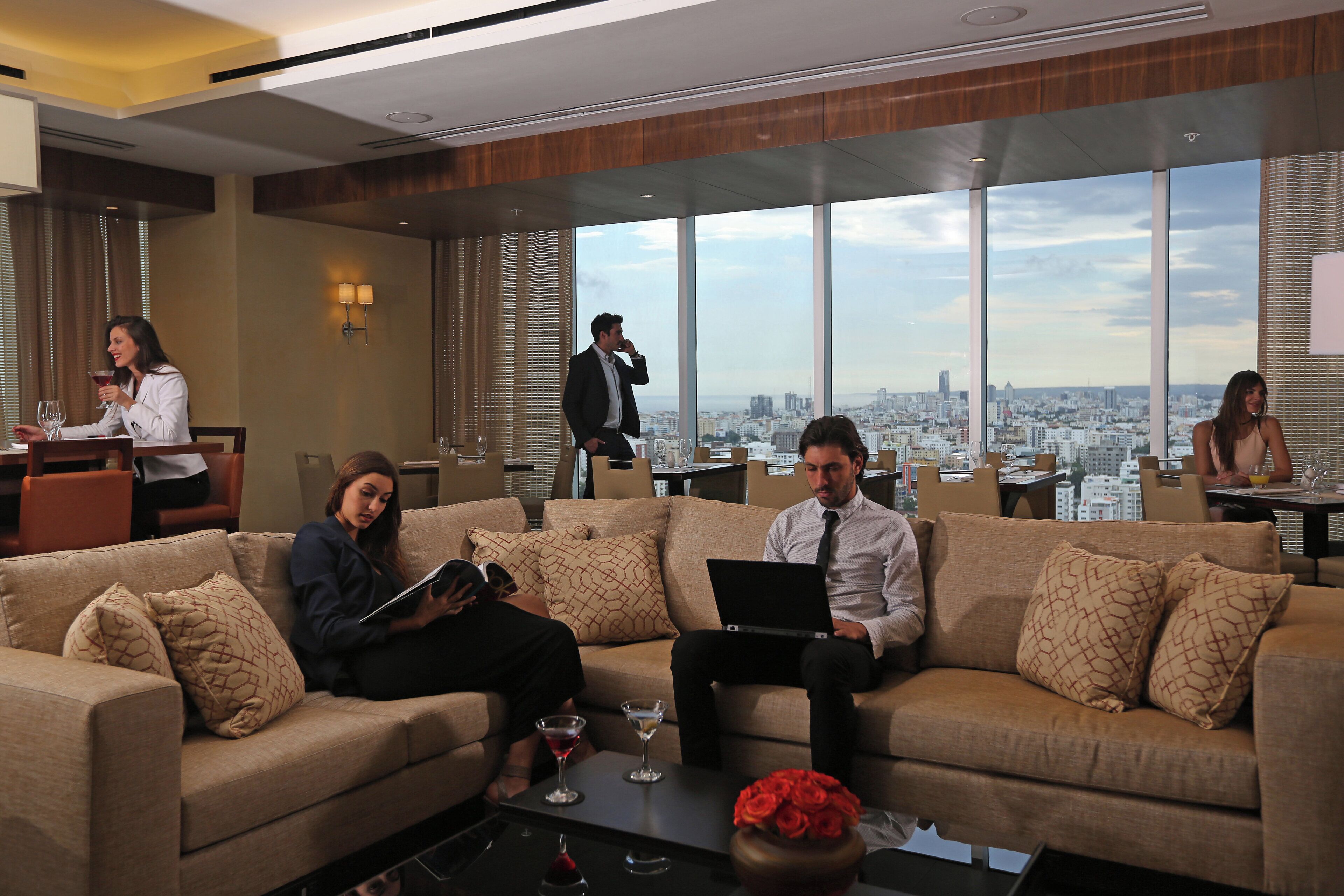 A modern hotel lobby with a large window offering a city view, a seating area with sofas, and a coffee table with a vase of flowers.