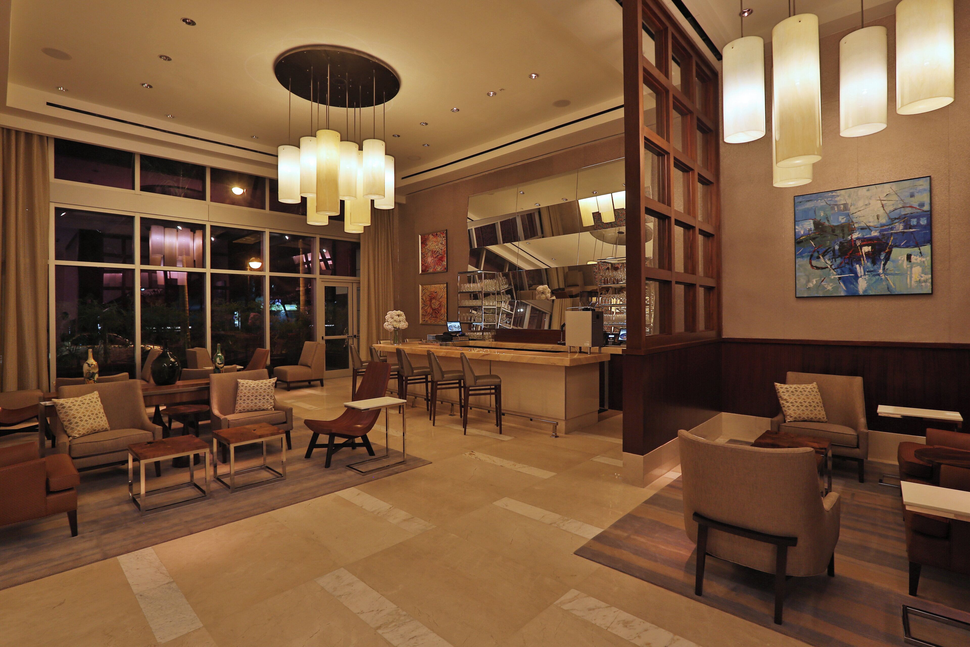 Un hall d’hôtel doté d’un bar, de différents espaces aménagés pour s’asseoir et de grandes fenêtres.