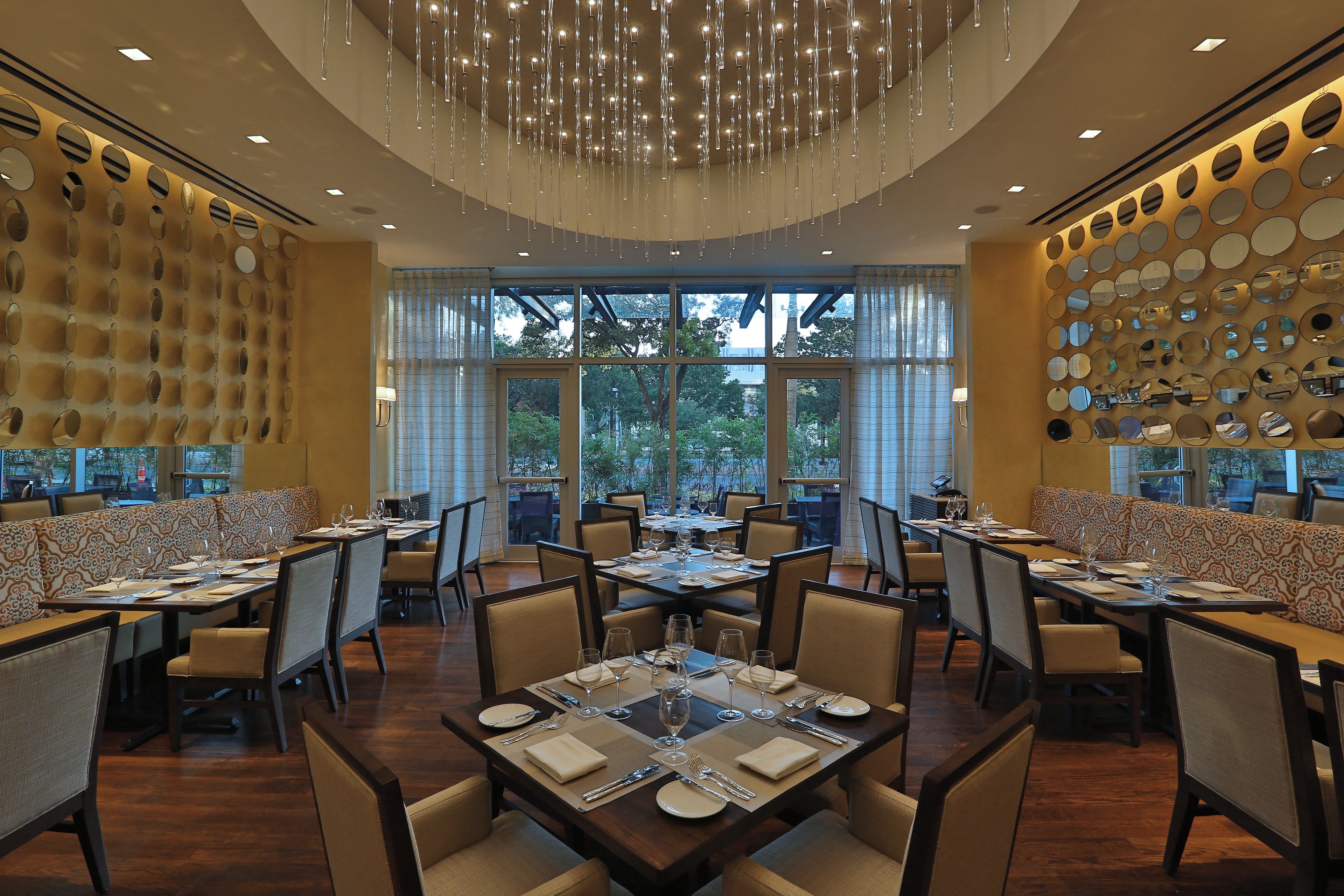 A restaurant with high ceilings, hanging light fixtures, and a wall of mirrors. Tables are set with white tablecloths and silverware.