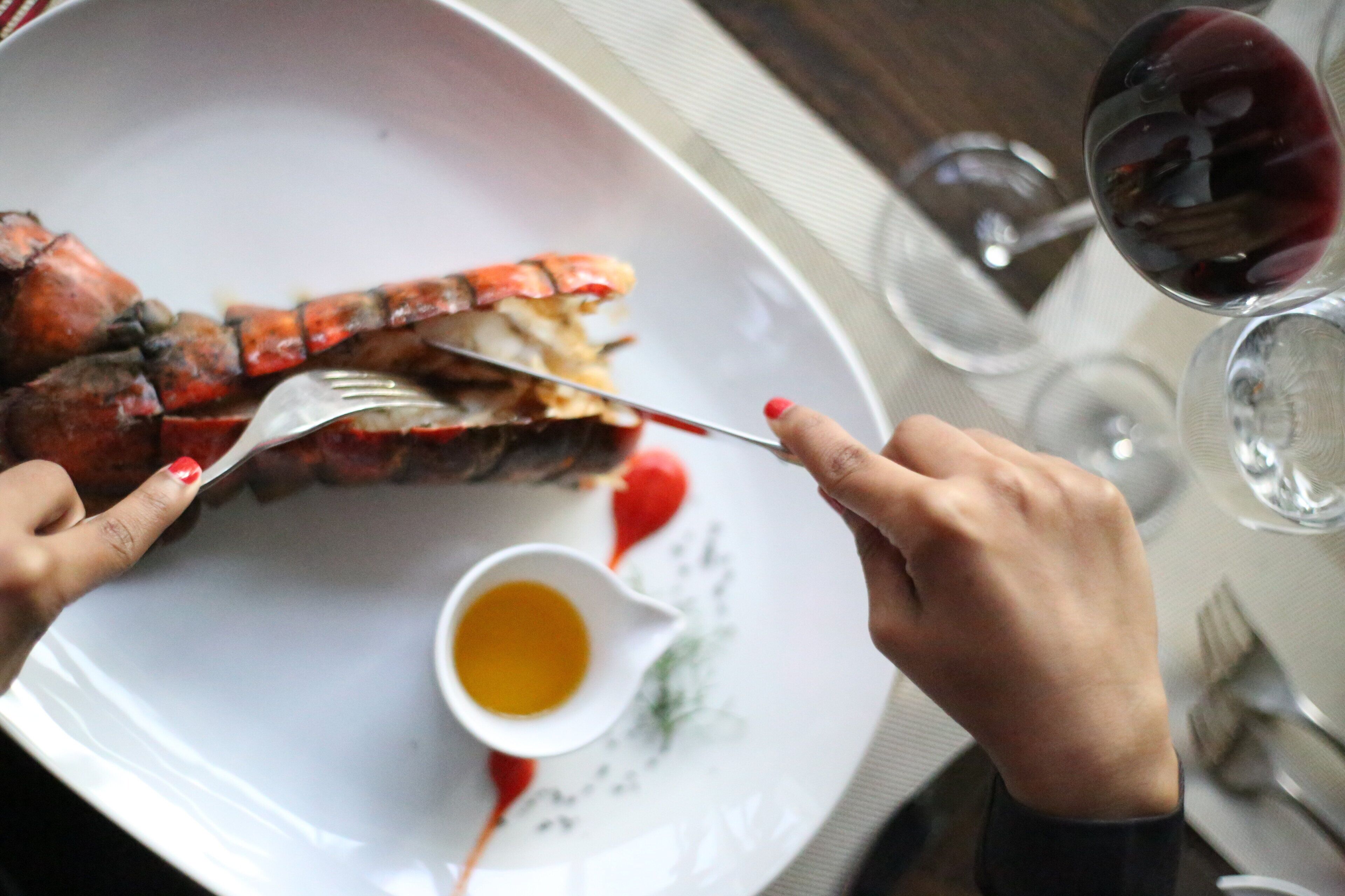 Un langostino siendo comido con tenedor y cuchillo, un pequeño plato de salsa y copas de vino tinto sobre una mesa.