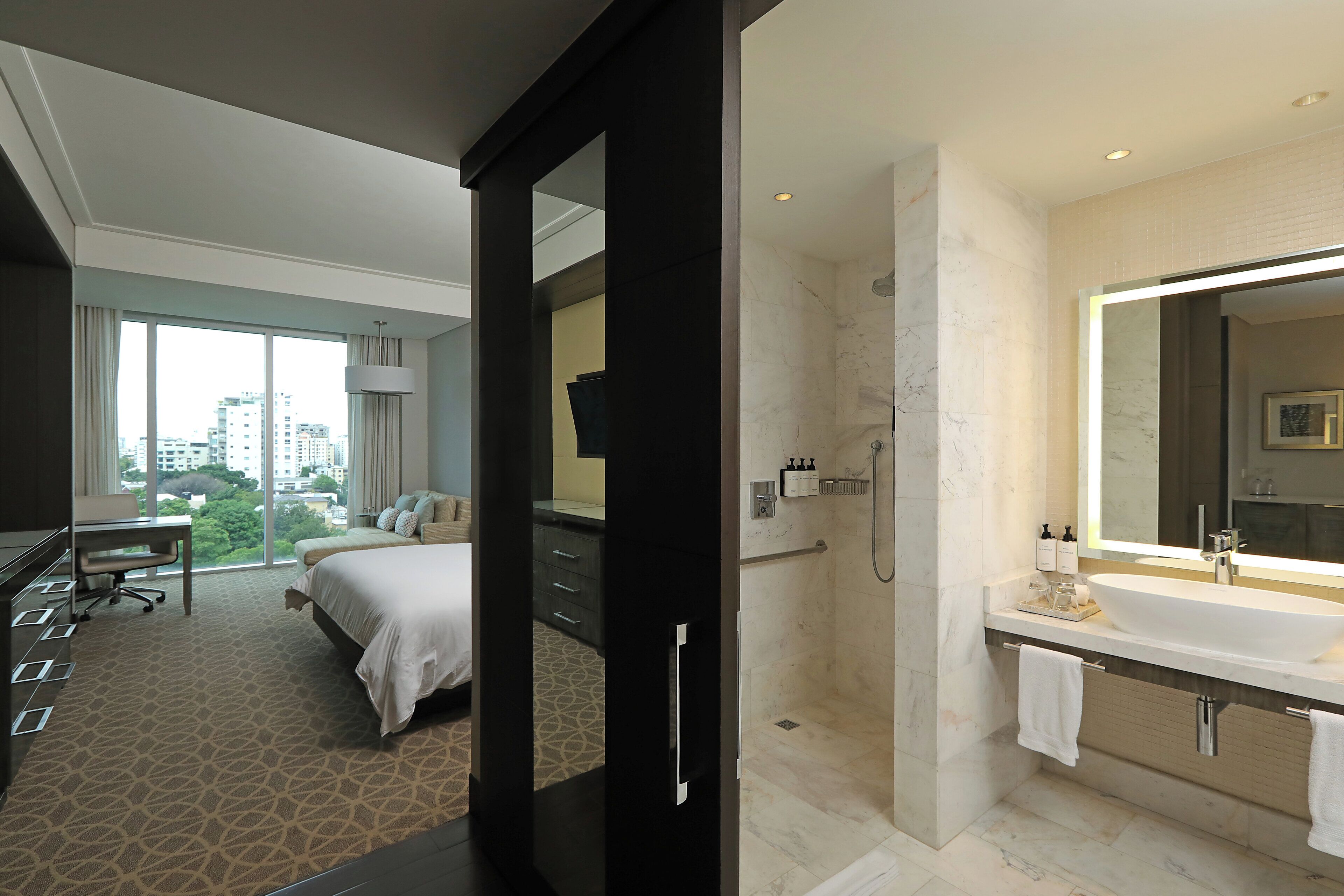 A hotel room with a large bed, a desk, and a view of the city. It includes a bathroom with a large mirror, sink, and shower.