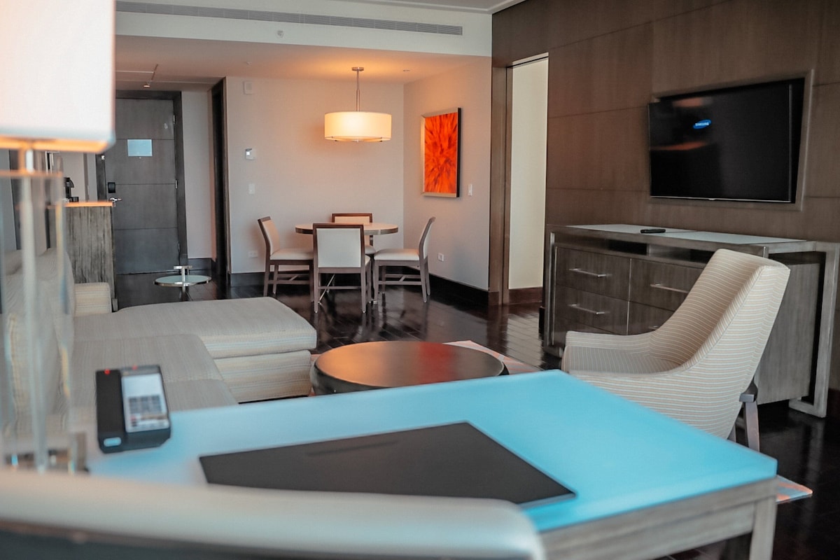 A hotel room with a bed, a desk, a chair, a TV, and a dining area.
