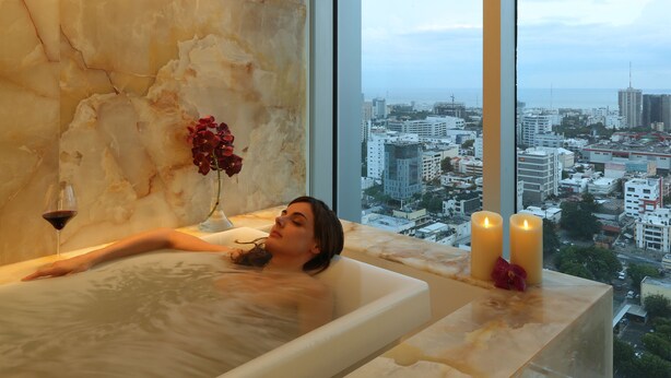 A person relaxing in a bathtub with a city view, a vase with flowers, and candles.
