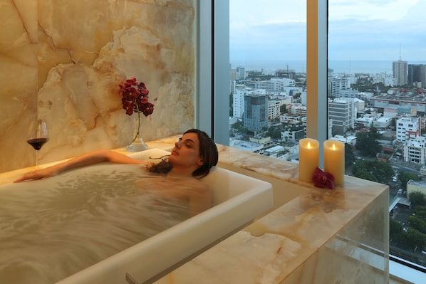 Una persona relajándose en una bañera con vistas a la ciudad, un florero con flores y velas.
