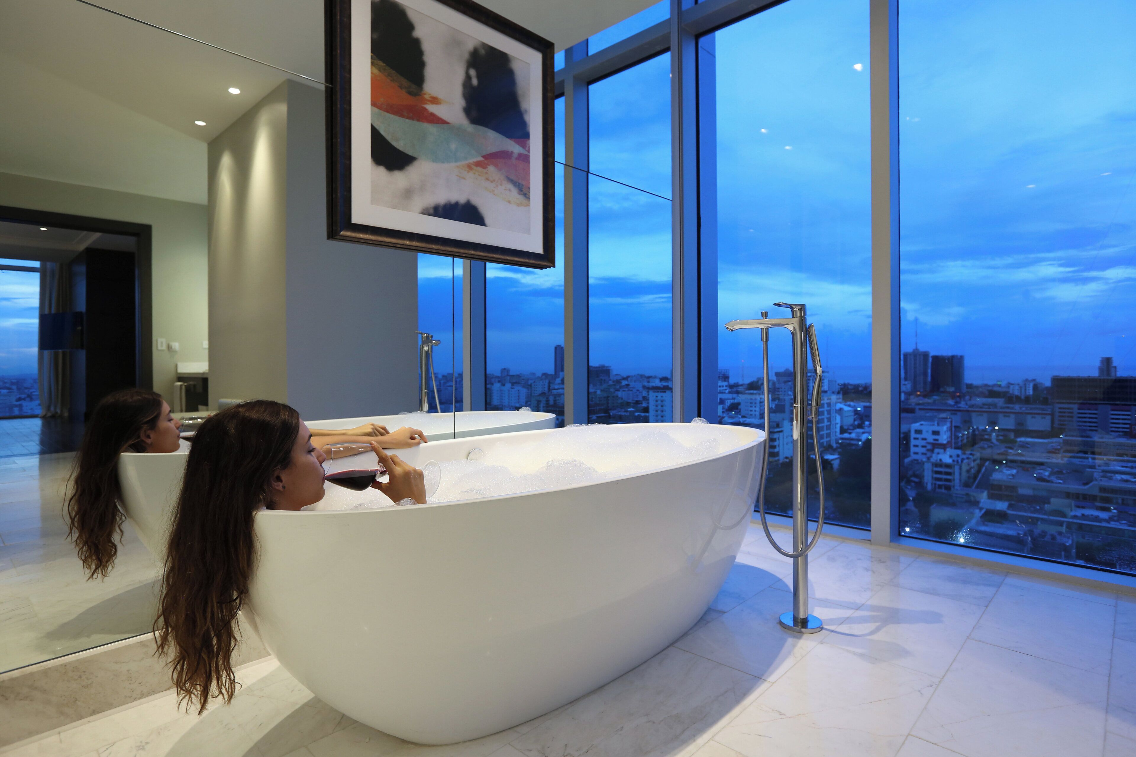 A modern bathroom with a large bathtub, a shower, and a city view through large windows.