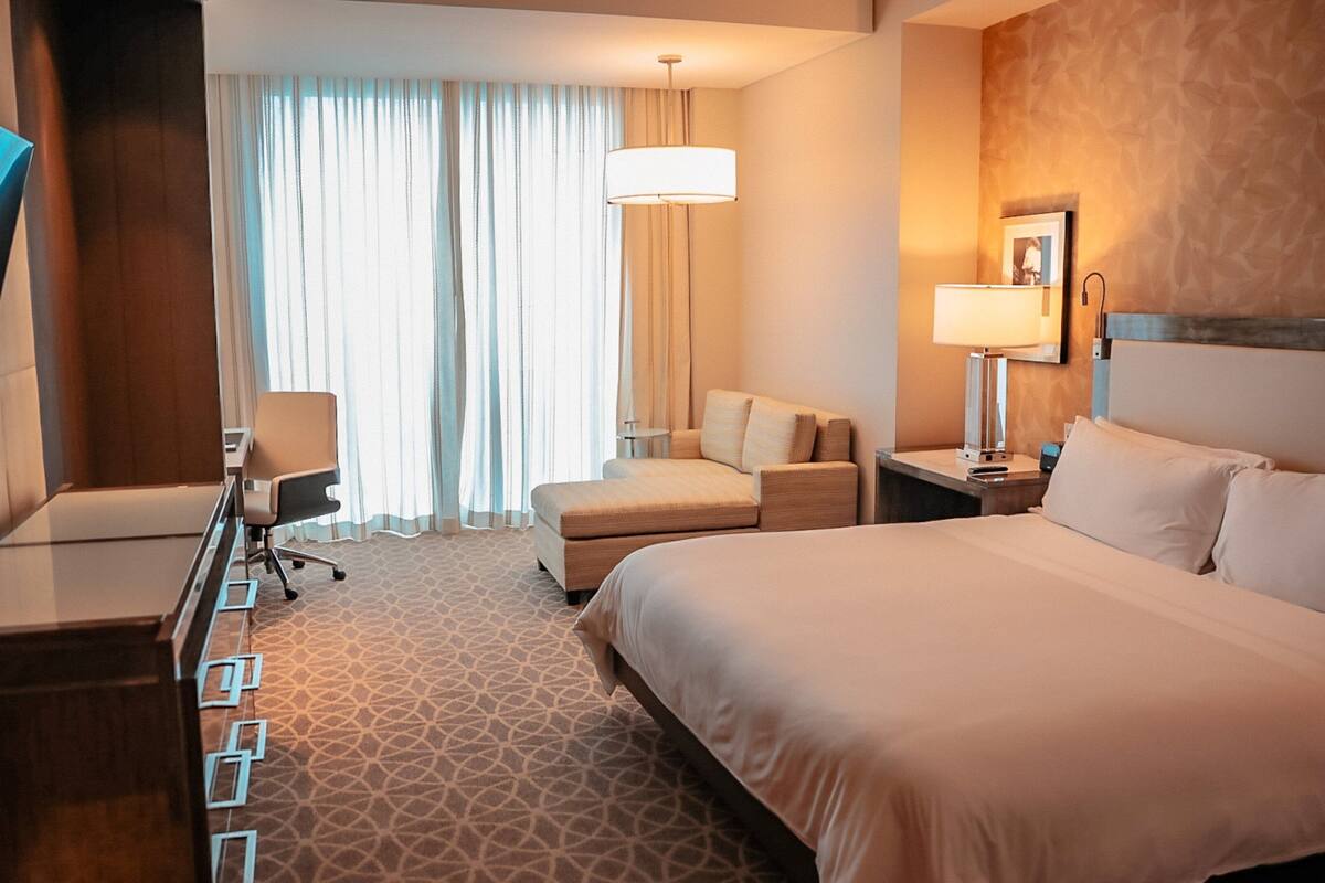 Ein Hotelzimmer mit Bett, Schreibtisch, Stuhl, Fernseher, Lampe und einem Fenster mit Vorhängen.
