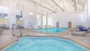 Ein Innenpool mit klarem Wasser, umgeben von Liegestühlen und Sonnenliegen, großen Fenstern und einer hohen Decke mit freiliegenden Leitungen.