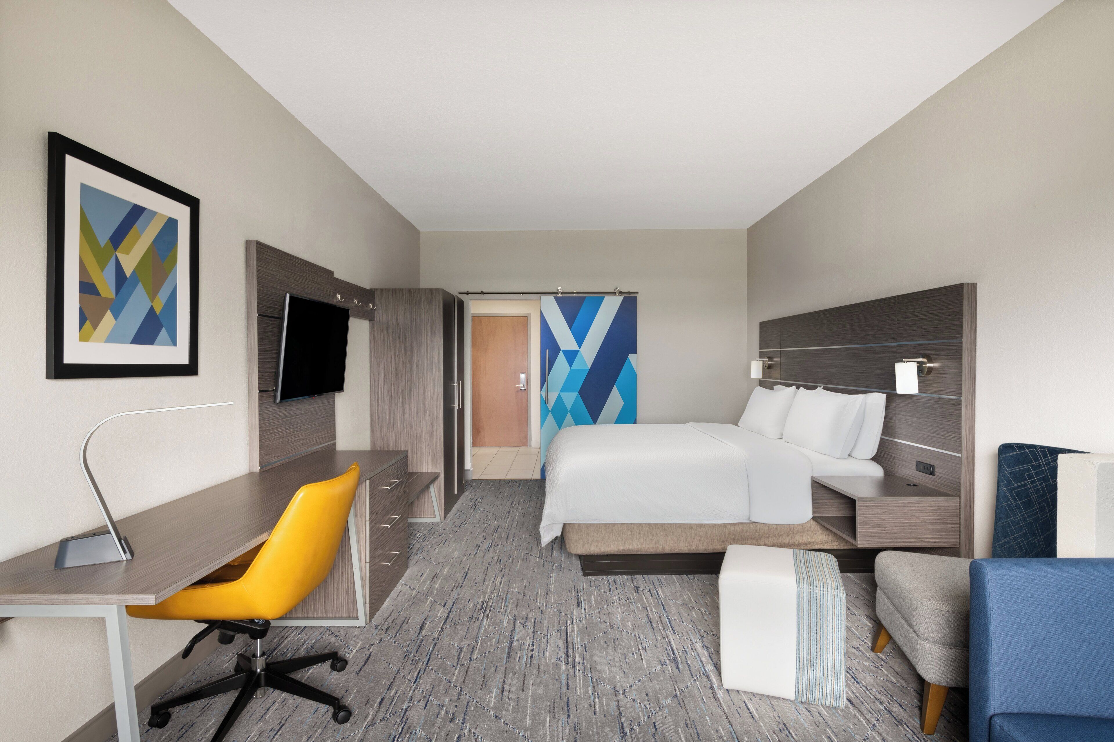 Quarto de hotel com cama, escrivaninha, cadeira, televisão e obras de arte nas paredes.