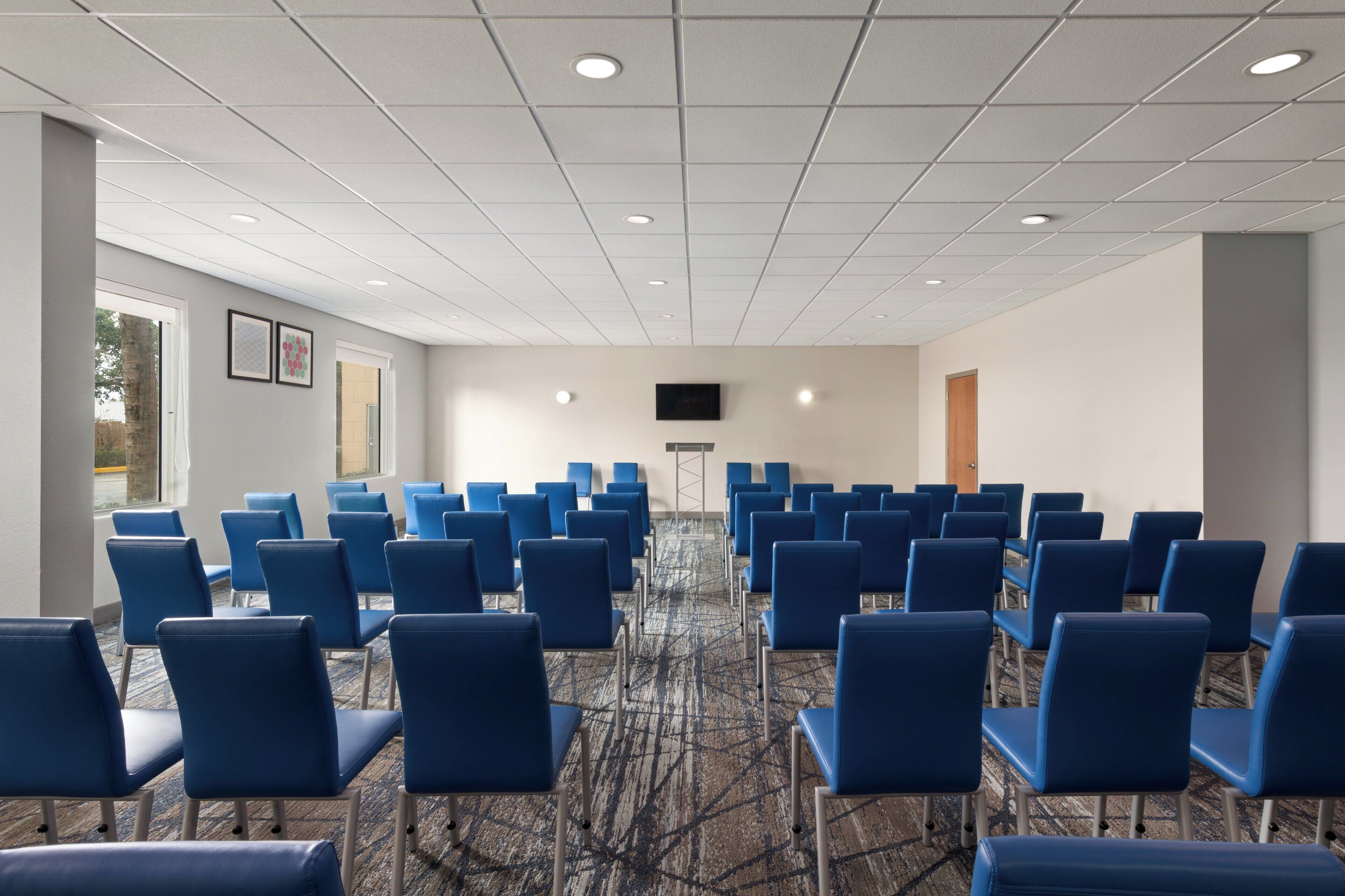 Un salón de conferencias con filas de sillas azules, suelo alfombrado y techo blanco con iluminación empotrada.