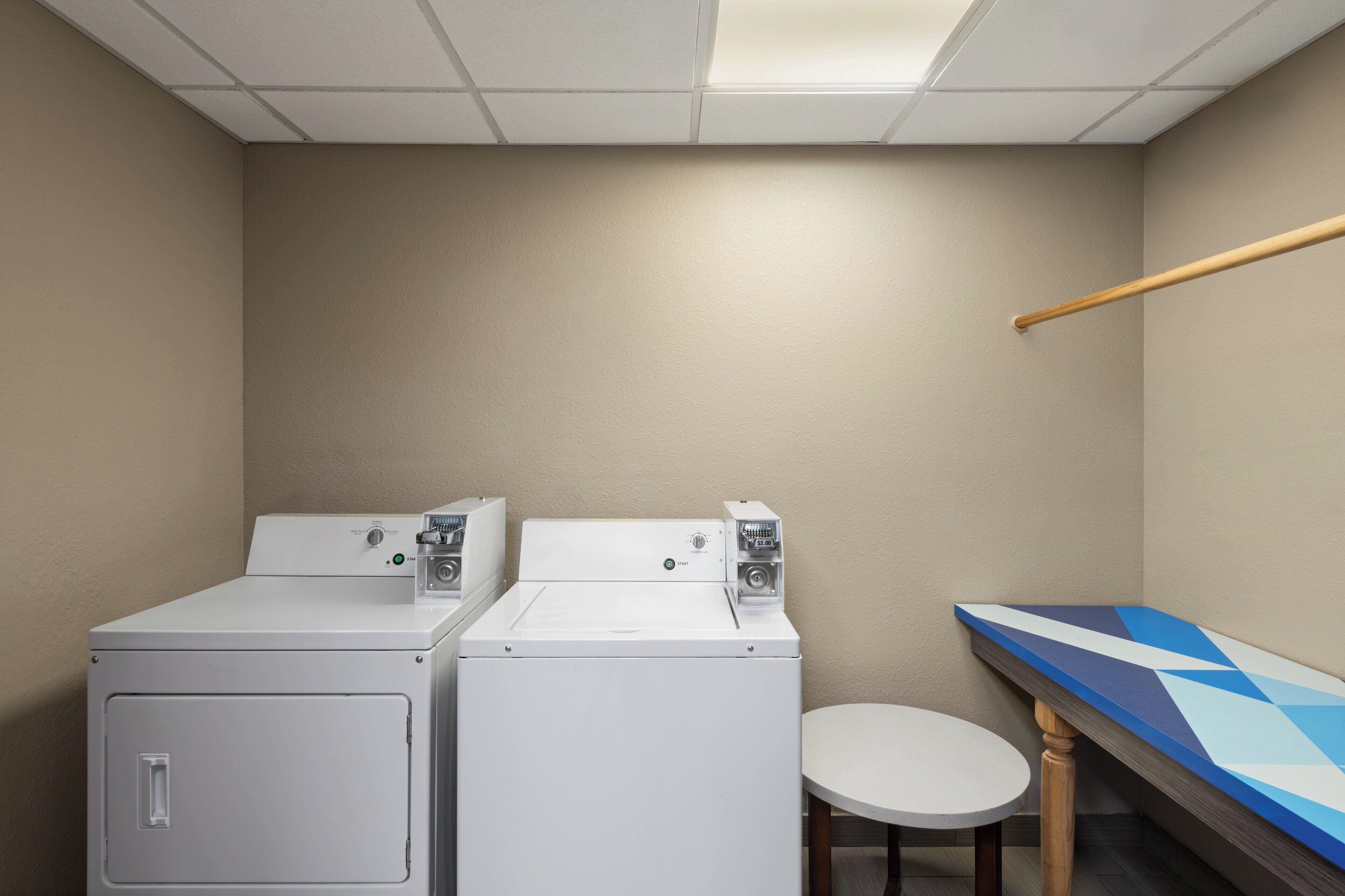 A laundry room with two white washers and dryers, a small round table, and a wooden bench with a geometric-patterned top.