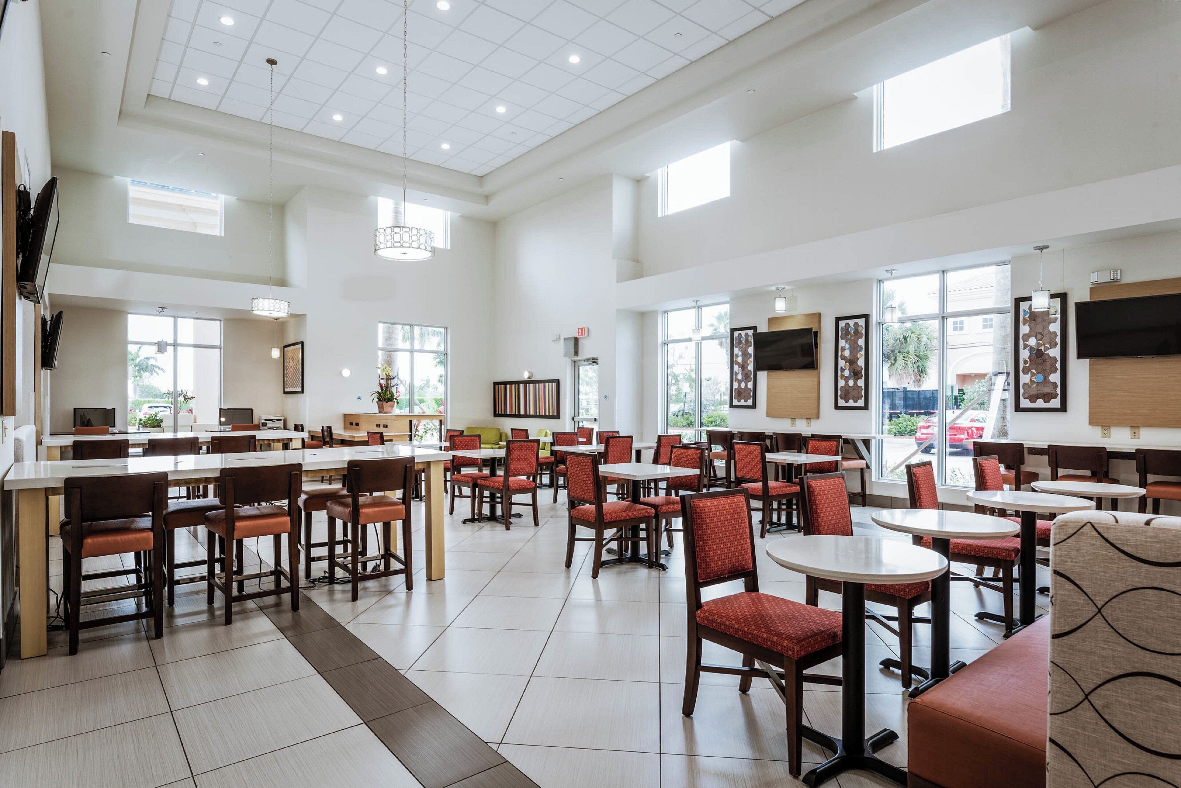 A spacious dining area with multiple tables and chairs, large windows, and a high ceiling with recessed lighting.