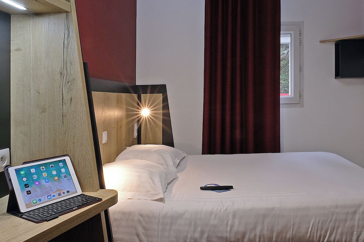 Une chambre d’hôtel avec un lit, un bureau avec un ordinateur portable, une télécommande et une fenêtre avec des rideaux.