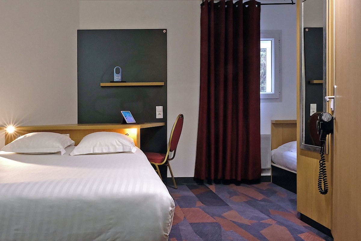 Une chambre d’hôtel avec un lit, un bureau avec un téléphone, une chaise et une fenêtre avec des rideaux.
