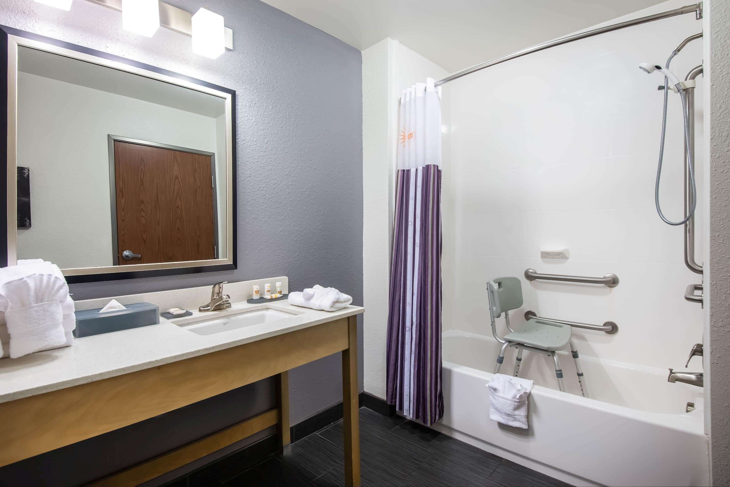 A bathroom with a double sink, a mirror, a shower with a grab bar, and a wheelchair-accessible chair.