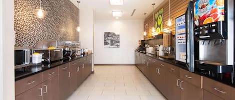 Une cuisine moderne avec des meubles de couleur brun foncé, un plan de travail noir et un sol carrelé. On y trouve un réfrigérateur avec un présentoir à fruits, une machine à café et un luminaire suspendu.
