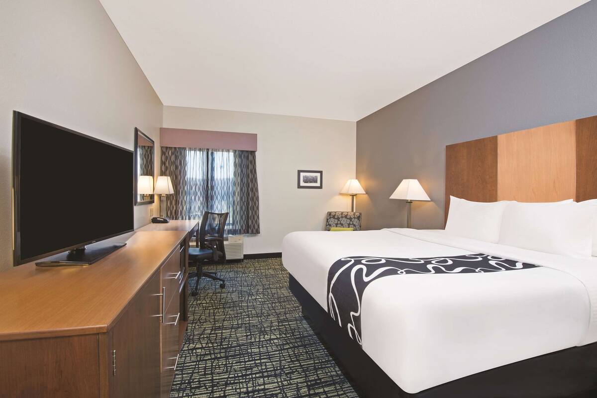 A hotel room with a bed, a desk with a TV, two lamps, a chair, and a patterned carpet.