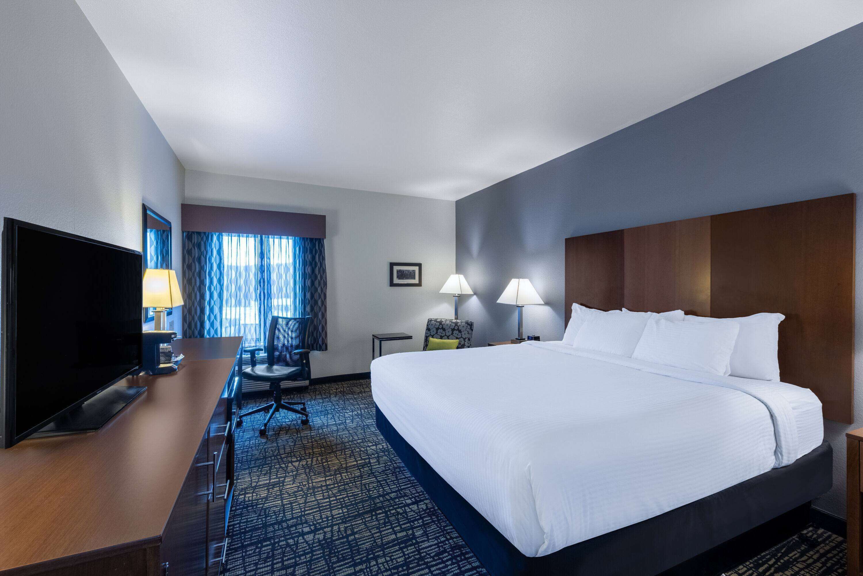 Una habitación de hotel con una cama grande, un escritorio con televisor, dos lámparas y una silla.
