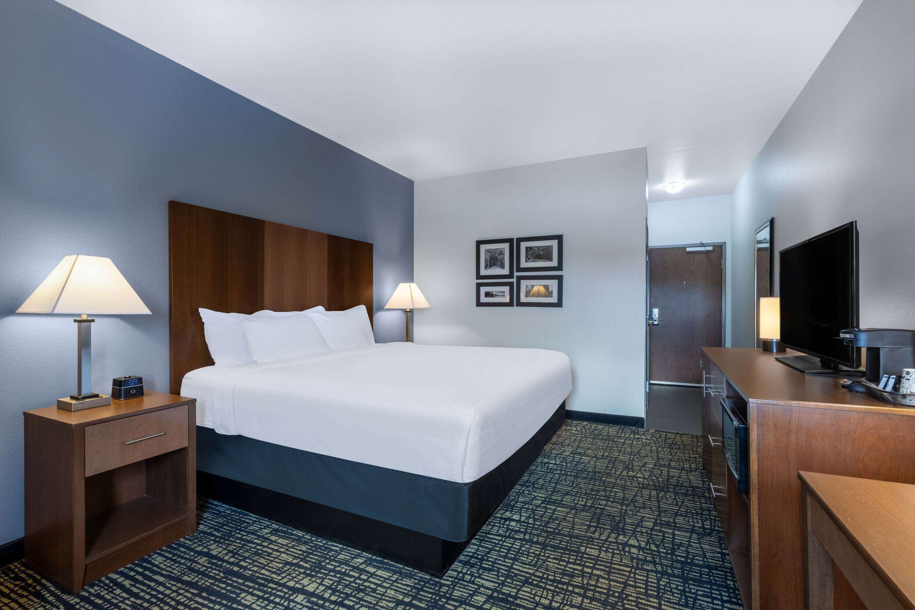 A hotel room with a large bed, two bedside tables with lamps, a flat-screen TV, a minibar, and a door leading to the bathroom.