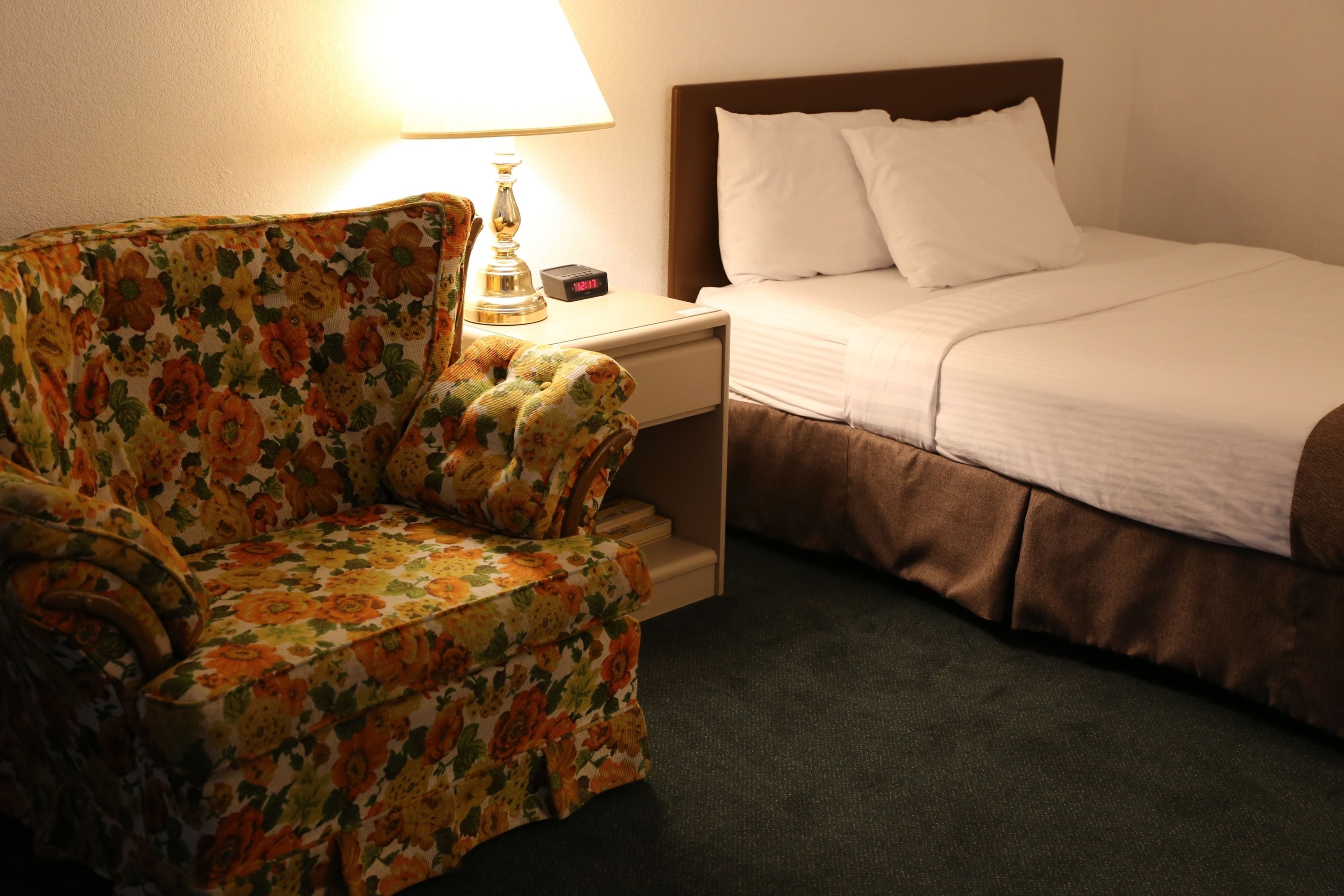 Una habitación de hotel con un sillón de estampado floral, una mesita de noche con lámpara y una cama bien hecha con ropa blanca.